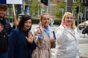 Na pierwszym planie trzy machające kobiety (ujęcie do pasa), w tle machający student. Wszyscy idą ulicami Szczecina w Spacerku na uniwerek. W tle kamienice i sygnalizacja świetlna.
