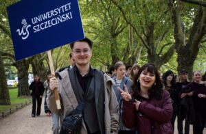 Park – aleja platanowa. Na pierwszym planie uśmiechnięty chłopak (po lewej) i dziewczyna (ujęcie do pasa). Chłopka trzyma granatowy transparent z białym napisem Uniwersytet Szczeciński. Dziewczyna pokazuje ręką znak Victorii. W tle młodzież – uczestnicy Spacerku na uniwerek.