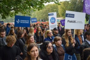 Młodzież idąca w pochodzie (około 40-osobowa grupa) – uczestnicy Spacerku na uniwerek. Niektórzy trzymają granatowe transparenty z białym napisem Uniwersytet Szczeciński.