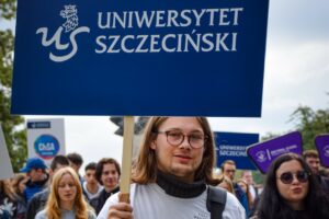 Na pierwszym planie student w okularach i długich włosach (ujęcie do piersi) trzyma granatowy transparent z białym napisem Uniwersytet Szczeciński. W tle młodzież – uczestnicy Spacerku na uniwerek.