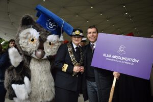 Po lewej stronie maskotka Uniwersytetu Szczecińskiego – sowa w granatowym birecie z białym logiem. W środku kobieta w mundurze Politechniki Morskiej. Po prawej przedstawiciel Klubu Absolwentów US trzymający fioletowy transparent z białym logiem klubu.