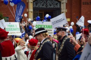 Uczestnicy Spacerku na uniwerek z flagami i transparentami, w tym władze uczelni ubrane w togi i mundury, mijają zabytkowy budynek rektoratu Uniwersytetu Szczecińskiego, przed którym stoją ludzie z białymi i granatowymi balonami. W tle transparent z napisem: Panie z dziekanatu – ogarniamy od 8:00.