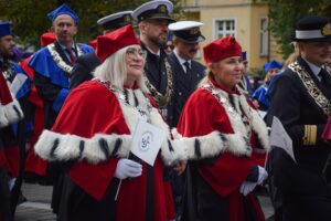 Idące w pochodzie dwie prorektorki Uniwersytetu Szczecińskiego w czerwonych togach z gronostajami. Trzymają małe białe papierowe flagi z granatowym logiem US. W tle władze innych uczelni w togach i mundurach.