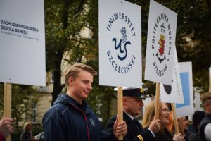 Uczestnicy Spacerku na uniwerek. Na pierwszym planie (ujęcie bokiem – do piersi) student w granatowej kurtce z białym logiem Uniwersytetu Szczecińskiego, trzymający transparent z logiem uczelni. Obok przedstawiciel Politechniki Morskiej w mundurze, trzymający swój transparent. Dalej przedstawiciele innych uczelni. W tle zielone drzewa.
