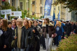 Uczestnicy Spacerku na uniwerek – wśród nich studenci ubrani w mundury Politechniki Morskiej. Uczestnicy trzymają różne transparenty i flagi. W tle zielone drzewa i kamienice.