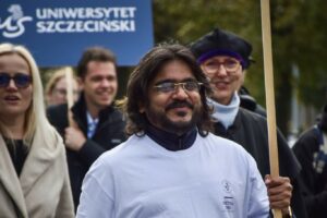 Uśmiechnięty mężczyzna w białej koszulce z logiem Uniwersytetu Szczecińskiego i Szkoły Doktorskiej US na piesi idzie w pochodzie podczas Spacerku na uniwerek. W tle inni uczestnicy, w tym jeden, który niesie granatowy transparent z białym logiem US.