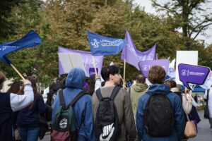 Uczestnicy Spacerku na uniwerek (ujęcie tyłem) machają granatowymi materiałowymi flagami z białym napisem Uniwersytet Szczeciński. Inni niosą fioletowe transparenty Szkoły Doktorskiej US.