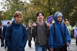 Na pierwszym planie trzech studentów. Chłopak w środku pokazuje obiema rękoma gest z uniesionym palcem wskazującym i małym nazywany „mano cornuta” lub „rogi”. W tle zielone drzewa i sygnalizacja świetlna oraz pozostali uczestnicy Spacerku na uniwerek.