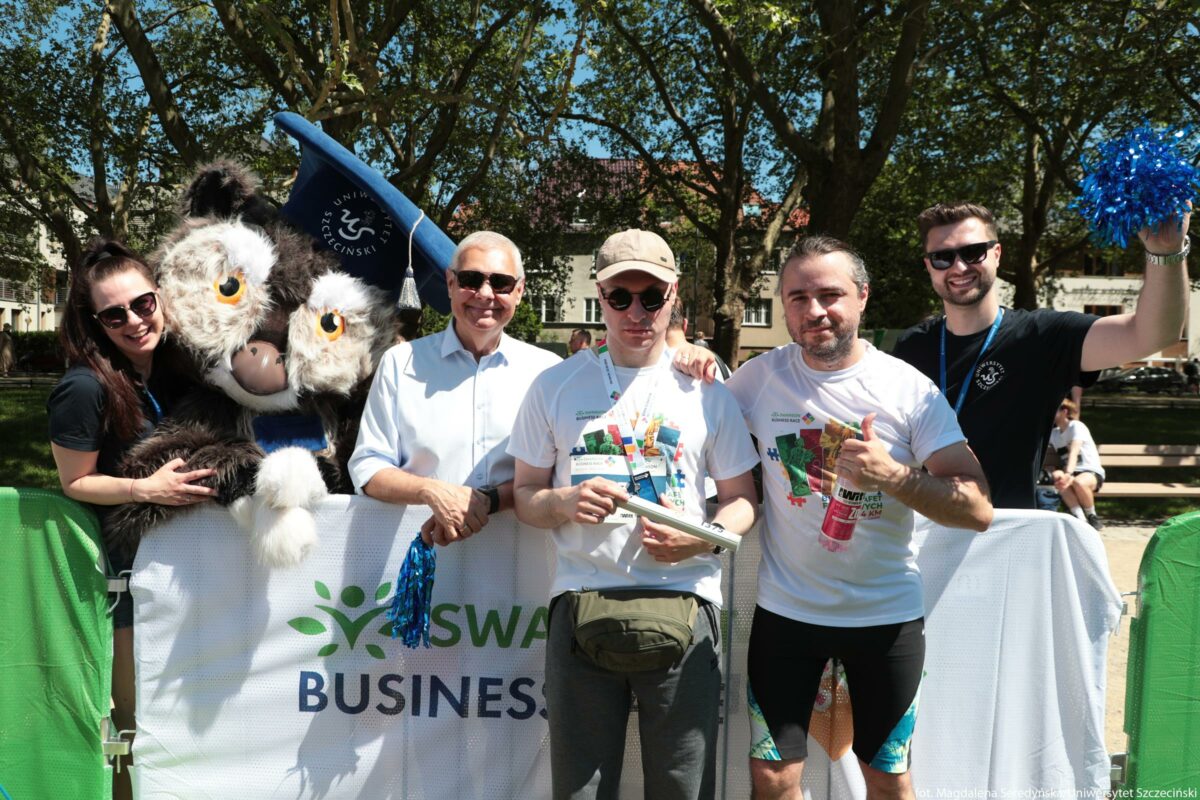Grupa sześciu osób stojąca na świeżym powietrzu przy białej barierce z logo „SWAN BUSINESS”. Po lewej stronie znajduje się wysoka pluszowa maskotka przypominająca sowę, w ciemnym futrze z dużymi żółtymi oczami i białymi policzkami; na głowie ma niebieski czworokątny biret z napisem „Uniwersytet Szczeciński”. Przytula się do niej młoda kobieta o jasnej skórze, z długimi ciemnymi włosami związanymi z tyłu; ma na sobie ciemną koszulkę i ciemne okulary przeciwsłoneczne, uśmiecha się w stronę aparatu. Obok stoi starszy mężczyzna o jasnej skórze, w jasnoniebieskiej koszuli z krótkim rękawem, również w ciemnych okularach przeciwsłonecznych; trzyma w dłoni niebieską błyszczącą pomponiastą ozdobę. W środku kadru stoi młody mężczyzna o jasnej skórze, w okrągłych okularach przeciwsłonecznych i beżowej czapce z daszkiem; ma na sobie białą koszulkę techniczną z kolorowym nadrukiem i smyczą z identyfikatorem na szyi, a w rękach trzyma biały rulon, przypominający pałeczkę sztafetową. Na biodrach ma oliwkową saszetkę. Po jego prawej stronie znajduje się mężczyzna o jasnej skórze, w białej sportowej koszulce z podobnym kolorowym nadrukiem i czarnych obcisłych spodenkach kolarskich z kolorowymi wstawkami na nogawkach; w jednej ręce trzyma różowy napój w butelce, drugą pokazuje uniesiony kciuk. Skrajnie po prawej stoi młody mężczyzna o jasnej skórze, w czarnej koszulce z małym jasnym logo na piersi i ciemnych okularach przeciwsłonecznych; na szyi ma niebieską smycz z identyfikatorem, a w uniesionej ręce trzyma niebieski pompon. W tle widoczne są drzewa z gęstą zieloną koroną i fragmenty budynków; pogoda jest słoneczna, cienie są wyraźne. Wszystkie osoby poza maskotką są uśmiechnięte i stoją blisko siebie, sprawiając wrażenie grupy uczestników wydarzenia lub imprezy plenerowej.