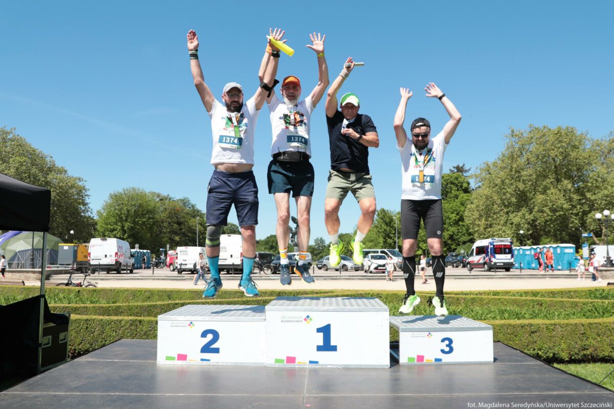 Czterech mężczyzn w trakcie skoku na podium biegowym na zewnątrz w słoneczny dzień. Stoją na trzech białych blokach podium oznaczonych kolejno: z lewej „2”, pośrodku „1”, z prawej „3”. Dwaj mężczyźni po lewej i mężczyzna po prawej mają na sobie białe koszulki techniczne z kolorowym nadrukiem z przodu, krótkie spodenki biegowe i numery startowe przypięte na brzuchu (1374 i 1174). Na nogach mają buty do biegania w żywych kolorach oraz ochraniacze lub opaski kompresyjne na łydkach i kolanach. Mężczyzna drugi od prawej ma czarną koszulkę, jasne krótkie spodenki i neonowożółte buty biegowe, w ręku trzyma pałeczkę sztafetową. Mężczyzna drugi od lewej również trzyma żółtą pałeczkę. Wszyscy są w powietrzu, ręce mają uniesione wysoko nad głowami w geście radości. Na szyjach wiszą im medale na taśmach. Na głowach trzech z nich są czapki z daszkiem w różnych kolorach, czwarty ma opaskę. W tle widoczna jest alejka parkowa z drzewami, zaparkowanymi samochodami dostawczymi i osobowymi oraz mobilnymi toaletami. Niebo jest bezchmurne i jasnoniebieskie, co sugeruje dobrą pogodę. Na podium po lewej i prawej stronie na froncie widać małe, kolorowe kwadratowe elementy graficzne i napisy, ale z tego ujęcia nie są one w pełni czytelne.