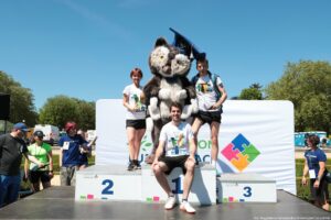 Podium z trzema miejscami na zewnątrz podczas imprezy sportowej. Na najwyższym, środkowym stopniu z numerem 1 siedzi młody mężczyzna o jasnej skórze, w białej koszulce z kolorowym nadrukiem, czarnych szortach i białych butach sportowych z czerwonym spodem. Za nim, na stopniu z numerem 2 po lewej, stoi młoda kobieta o jasnej skórze i krótkich rudawych włosach, w białej koszulce z tym samym nadrukiem, czarnych szortach i ciemnych butach sportowych. Po prawej, na stopniu z numerem 3, stoi młody mężczyzna o jasnej skórze, w białej koszulce z takim samym nadrukiem, czarnych krótkich spodenkach, białych butach i z plecakiem na ramionach. Za trójką osób stoi duża maskotka w kostiumie puchatego zwierzęcia, szaro‑białego z dużymi uszami, w granatowej czapce absolwenckiej z jasnym chwostem. W tle widoczny jest biały baner z kolorowym logo złożonym z czterech elementów puzzli w kolorach niebieskim, różowym, żółtym i zielonym oraz fragment zielonego napisu. Po bokach podium stoją inne osoby w sportowych strojach i koszulkach organizatorów, a za nimi znajdują się drzewa, niebo bez chmur i pojedyncze pojazdy.