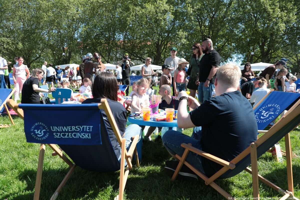 Plenerowy piknik rodzinny na trawie w słoneczny dzień. Na pierwszym planie stoją dwa granatowe leżaki z jasnym drewnianym stelażem, z nadrukiem „UNIWERSYTET SZCZECIŃSKI” i okrągłym logo uczelni po lewej stronie oparcia. Między leżakami znajduje się mały, okrągły, niebieski stolik, przy którym siedzi kilkoro dzieci; na stoliku leżą kolorowe przybory plastyczne, kartki i kubki z napojami w pomarańczowych, przezroczystych kubkach. Z lewej strony stolika siedzi dorosła osoba w granatowej koszulce z długimi, ciemnymi włosami, odwrócona plecami. Po prawej stronie, blisko aparatu, siedzi dorosły mężczyzna o jasnych włosach, w granatowej koszulce i dżinsach, pochylony do przodu nad stolikiem. Przy stoliku widać kilkoro małych dzieci w jasnych ubraniach; jedno dziecko ma jasnoróżową bluzkę i różowe, odblaskowe okulary przeciwsłoneczne. W tle, na trawie, rozstawione są kolejne leżaki i stoliki, wokół których stoją i siedzą liczne osoby dorosłe oraz dzieci, część z nich w letnich kapeluszach i okularach przeciwsłonecznych. Jeszcze dalej widać białe namioty lub stoiska oraz osoby spacerujące i rozmawiające; pośrodku stoją dwie osoby w strojach przypominających zbroje rycerskie. Cała scena odbywa się w parku; w tle rośnie rząd gęstych, zielonych drzew pod jasnoniebieskim, bezchmurnym niebem.