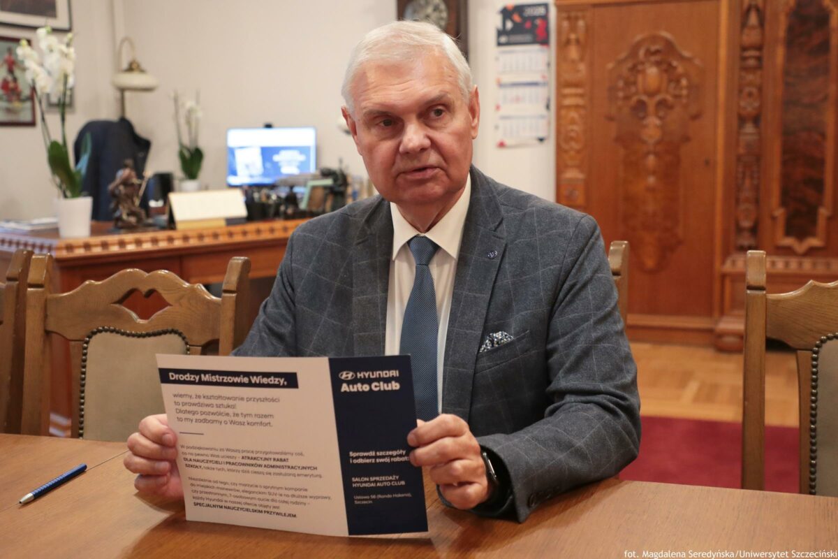 Starszy mężczyzna o jasnej skórze i krótkich siwych włosach siedzący przy drewnianym stole i trzymający rozłożoną ulotkę. Ma na sobie szary garnitur w kratę, białą koszulę i niebieski krawat; patrzy w bok z poważnym wyrazem twarzy. Na ulotce widać m.in. nagłówek „Drodzy Mistrzowie Wiedzy,” oraz logo i tekst „HYUNDAI Auto Club”; reszta tekstu jest drobna i częściowo nieczytelna w tej jakości. Na stole leży długopis. Tło przypomina gabinet: biurko z komputerem, rośliny doniczkowe, kalendarz na ścianie oraz duże, ozdobnie rzeźbione drewniane drzwi. W prawym dolnym rogu zdjęcia jest podpis: „fot. Magdalena Seredyńska / Uniwersytet Szczeciński”.