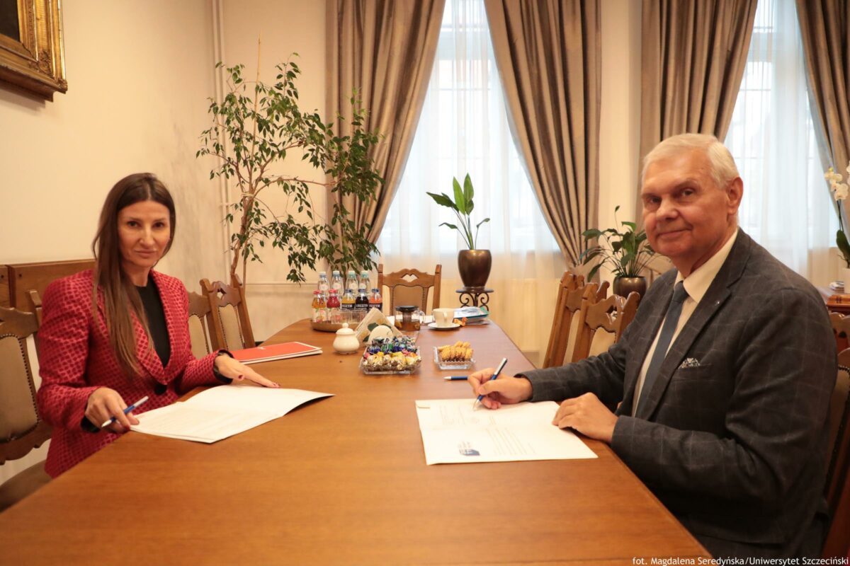 Dwie osoby siedzące przy długim drewnianym stole w gabinecie i podpisujące dokumenty. Po lewej stronie siedzi kobieta o jasnej skórze, z długimi prostymi brązowymi włosami, w czerwonej marynarce we wzór i czarnej bluzce; trzyma długopis nad rozłożonymi kartkami. Po prawej stronie siedzi starszy mężczyzna o jasnej skórze, z krótkimi siwymi włosami, w ciemnej marynarce, białej koszuli i jasnym krawacie; również trzyma długopis nad dokumentem. Na stole stoją butelki z napojami, cukiernica, patera z cukierkami i małe przekąski na talerzyku. W tle są wysokie okna z jasnymi firanami i beżowymi zasłonami, kilka roślin doniczkowych oraz drewniane krzesła. W prawym dolnym rogu widoczny jest podpis: „fot. Magdalena Seredyńska/Uniwersytet Szczeciński”.