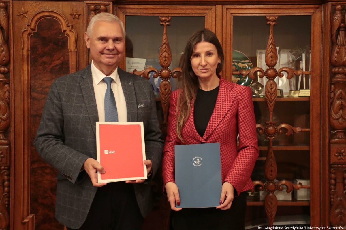 Dwie osoby stojące obok siebie na tle dużej, rzeźbionej, drewnianej witryny z przeszklonymi drzwiami. Po lewej stoi starszy mężczyzna o jasnej karnacji, z krótkimi siwymi włosami, ubrany w szarą marynarkę w kratę, białą koszulę i jasnoniebieski krawat. Trzyma czerwoną teczkę z białą ramką; na niej widać małe białe logo i napis „GRUPA ECHO”. Po prawej stoi kobieta o jasnej karnacji, z długimi brązowymi włosami, ubrana w czerwony żakiet we wzór i czarną bluzkę. Trzyma granatową teczkę z białym godłem i napisem „UNIWERSYTET SZCZECIŃSKI”. W witrynie za nimi widać różne przedmioty i pamiątki, w tym dyplomy i dekoracyjne elementy. W prawym dolnym rogu zdjęcia jest podpis: „fot. Magdalena Seredyńska/Uniwersytet Szczeciński”.