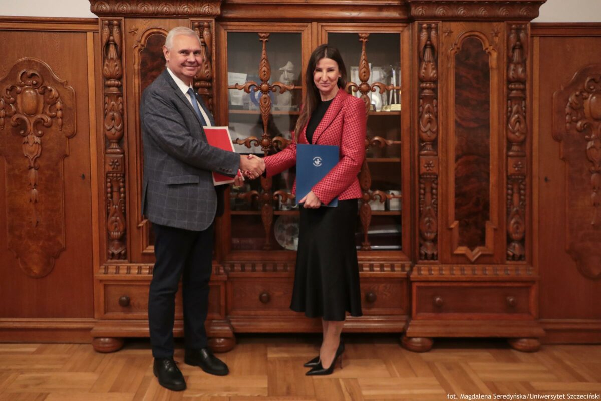 Dwie osoby stojące w pomieszczeniu przed dużą, rzeźbioną, drewnianą szafą z przeszklonymi drzwiami. Po lewej stronie stoi starszy mężczyzna o jasnej skórze i siwych, krótko przyciętych włosach. Ma na sobie szarą marynarkę w kratę, białą koszulę i ciemny krawat oraz ciemne spodnie i czarne buty. Trzyma w lewej ręce czerwoną teczkę. Po prawej stronie stoi kobieta o jasnej skórze i długich, ciemnych włosach. Ma na sobie czerwony żakiet w drobny wzór, czarną bluzkę i czarną spódnicę do połowy łydki oraz czarne szpilki. Trzyma niebieską teczkę z białym godłem lub logo. Osoby podają sobie dłonie na środku kadru. Na dole po prawej stronie widoczny jest napis: „fot. Magdalena Seredyńska/Uniwersytet Szczeciński”.