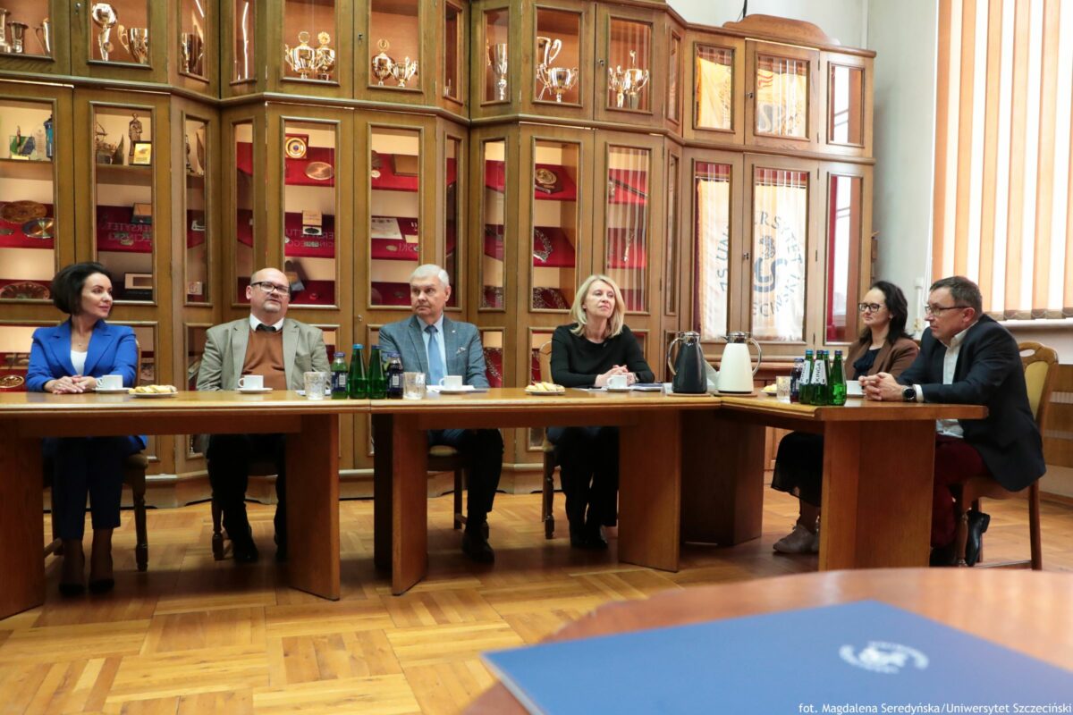 Długi drewniany stół konferencyjny w sali z dużymi, przeszklonymi gablotami w tle, wypełnionymi pucharami i innymi eksponatami. Przy stole siedzi sześć osób, ustawionych w jednym rzędzie. Od lewej: kobieta o jasnej skórze w niebieskim garniturze. Obok mężczyzna o jasnej skórze w okularach, w jasnej marynarce i brązowym swetrze. Dalej mężczyzna o jasnej skórze z siwymi włosami w niebieskim garniturze i krawacie. Następnie kobieta o jasnej skórze z jasnymi włosami, w czarnym stroju. Po prawej stronie siedzi kobieta o jasnej skórze w okularach, w ciemnym stroju, a skrajnie po prawej mężczyzna o jasnej skórze w okularach, w ciemnej marynarce i białej koszuli. Na stole stoją filiżanki, szklanki, talerzyki z ciastkami, butelki wody oraz dwa termosy/dzbanki (jeden ciemny, drugi biały). Po prawej widać kilka zielonych butelek z etykietami ustawionych obok siebie. Na pierwszym planie, u dołu zdjęcia, częściowo widoczna jest niebieska teczka lub okładka z jasnym znakiem graficznym. W prawym tle są wysokie okna z pionowymi żaluzjami. W prawym dolnym rogu widnieje podpis: „fot. Magdalena Seredyńska/Uniwersytet Szczeciński”.