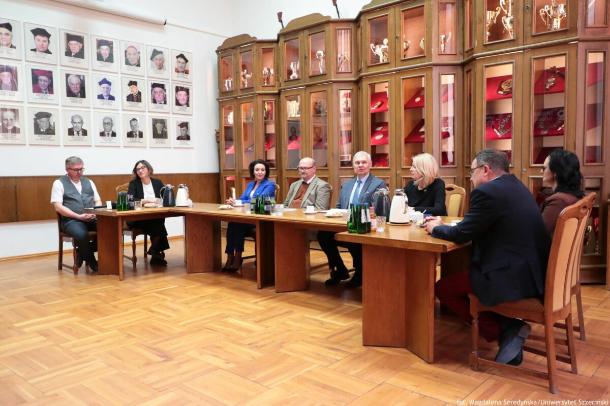 Sala konferencyjna z długim, drewnianym stołem ustawionym w kształcie podkowy oraz grupą siedmiu osób w trakcie spotkania. Podłoga jest z jasnego parkietu, a ściany częściowo wyłożone drewnianą boazerią. Po lewej stronie na ścianie wisi wiele oprawionych portretów (czarno-białe i kolorowe) ułożonych w równych rzędach; pod każdym zdjęciem znajduje się podpis, ale tekst jest zbyt mały, żeby go odczytać. W tle po prawej stoją wysokie, drewniane gabloty z przeszklonymi drzwiczkami; wewnątrz są puchary, małe statuetki i czerwone ekspozytory z medalami lub odznaczeniami. Przy stole siedzą osoby w strojach formalnych. Od lewej: mężczyzna w białej koszulce i szarej kamizelce; obok kobieta w ciemnym stroju i okularach; dalej kobieta w niebieskim stroju; mężczyzna w jasnej marynarce i okularach; mężczyzna w garniturze; kobieta z jasnymi włosami w ciemnym stroju; na pierwszym planie z prawej mężczyzna w ciemnej marynarce, obok niego kobieta z ciemnymi włosami. Na stole stoją zielone butelki wody, szklanki, dzbanki/termosy oraz drobne notatki lub kartki. W prawym dolnym rogu widoczny jest napis kredytowy: „fot. Magdalena Seredyńska/Uniwersytet Szczeciński”.