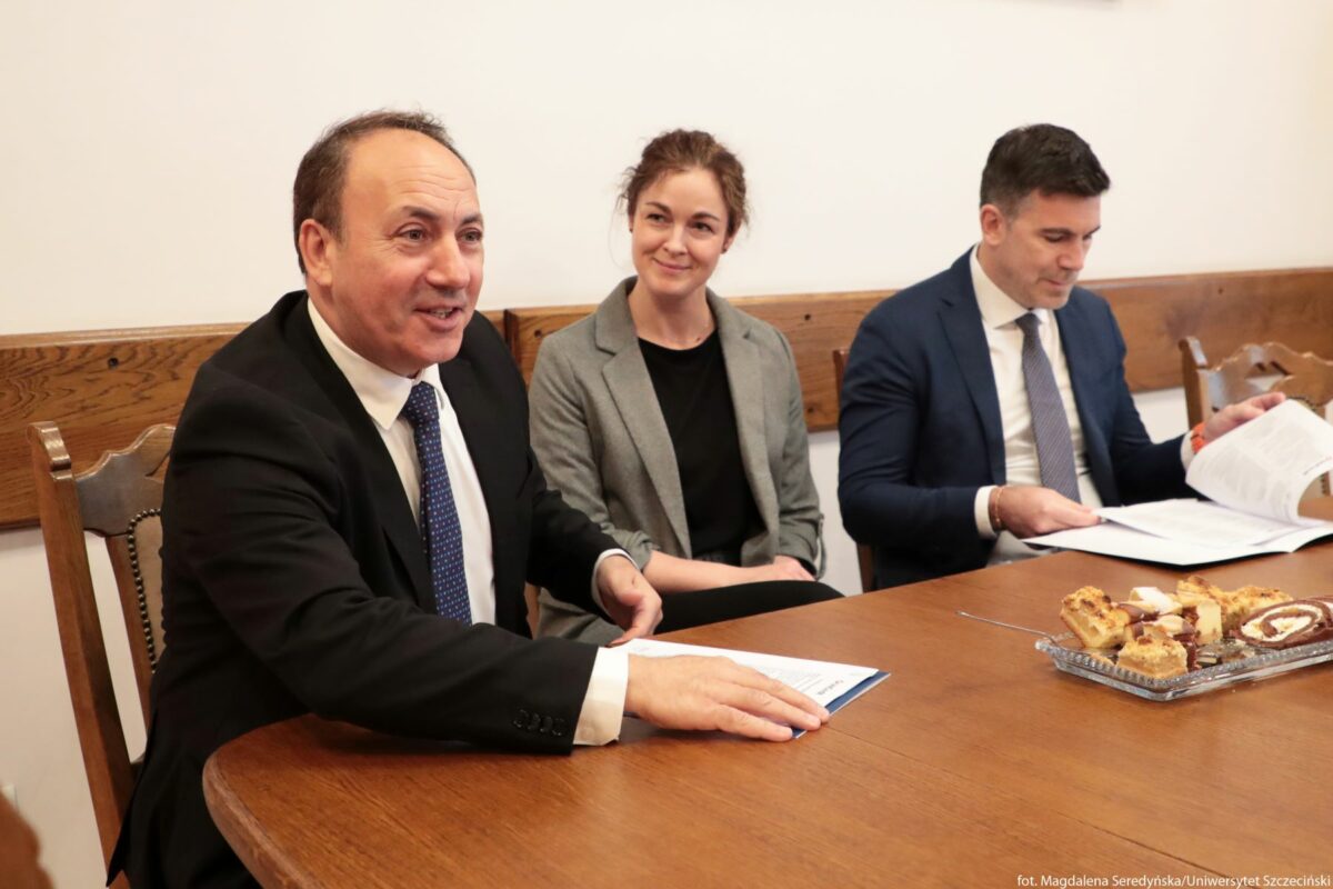 Trzy osoby siedzące przy drewnianym stole w sali spotkań. Po lewej stronie znajduje się mężczyzna w średnim wieku o jasnej karnacji, z krótkimi, przerzedzonymi, ciemnymi włosami. Ma na sobie czarną marynarkę, białą koszulę i granatowy krawat w drobny wzór. Jest zwrócony lekko w stronę aparatu, uśmiecha się i ma przed sobą kartkę papieru, na której opiera dłonie. Na środku siedzi młoda kobieta o jasnej karnacji i brązowych, upiętych włosach. Nosi szarą marynarkę i czarną bluzkę. Patrzy w stronę mężczyzny po lewej z lekkim uśmiechem, ma splecione dłonie ułożone na kolanach. Po prawej stronie siedzi mężczyzna o jasnej karnacji i ciemnych, krótko przyciętych włosach. Ma na sobie granatową marynarkę, białą koszulę i jasnoszary krawat. Skupia wzrok na dokumentach, które trzyma rozłożone przed sobą nad stołem. Przed osobami stoi prostokątny drewniany stół. Na jego brzegu, bliżej prawej strony, znajduje się szklany półmisek z kilkoma kawałkami ciasta o jasnobrązowym kolorze, w tym roladą z jasnym nadzieniem. W tle widoczne są drewniane oparcia krzeseł i fragment drewnianej listwy przy ścianie w jasnym pomieszczeniu. Na dole, w prawym rogu, znajduje się mały biały napis z informacją o autorce zdjęcia i nazwie uczelni.