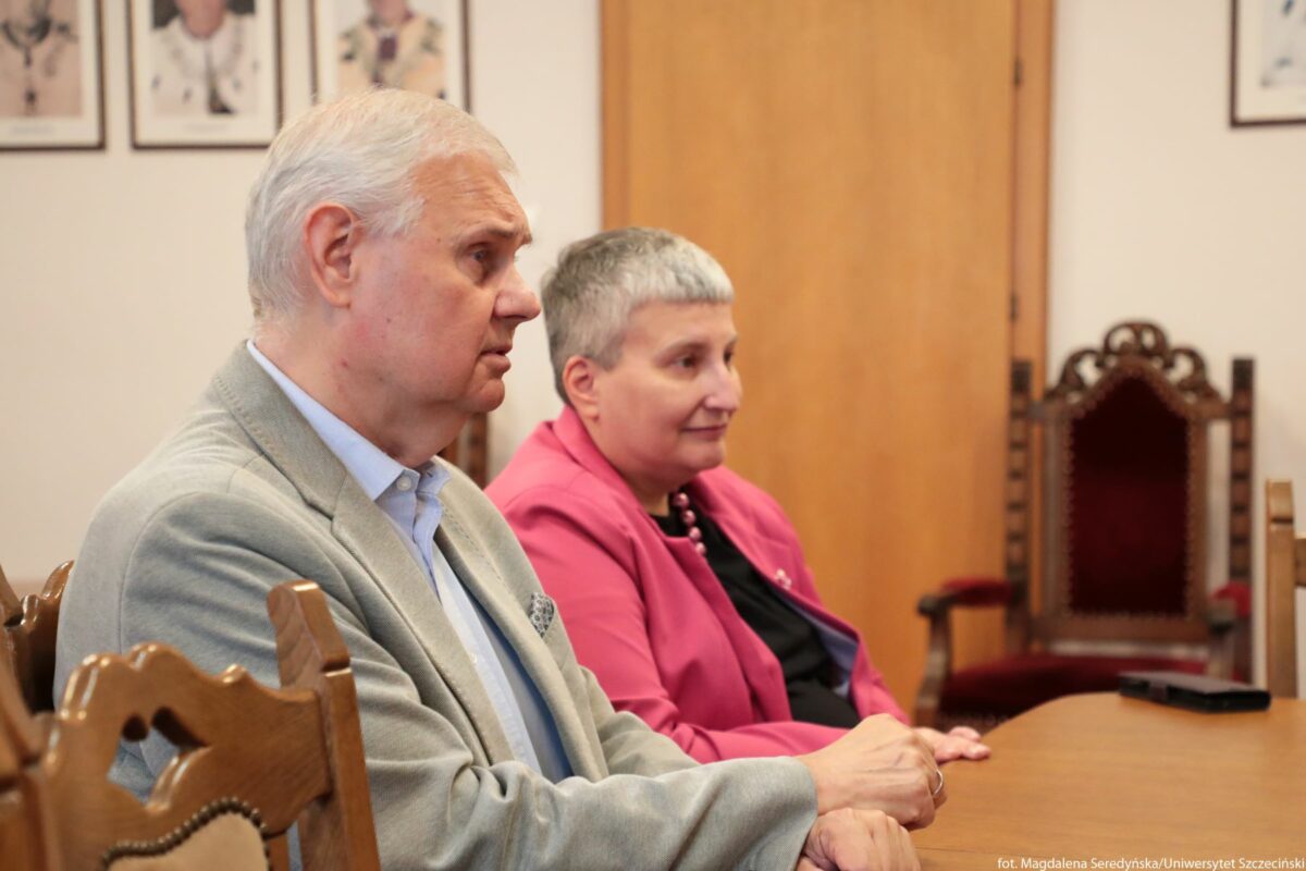 Dwoje starszych dorosłych siedzących przy drewnianym stole w sali o urzędowym charakterze. Po lewej stronie znajduje się mężczyzna o jasnej, siwiejącej fryzurze, jasnej karnacji i pełniejszej twarzy. Ma na sobie jasnoszarą marynarkę i jasnoniebieską koszulę z kołnierzykiem, w kieszonce marynarki widać poszetkę. Jego ręce są złożone przed sobą na krawędzi stołu, ma poważny, skupiony wyraz twarzy i patrzy w prawo, poza kadr. Po prawej stronie siedzi kobieta o krótkich, bardzo krótko ostrzyżonych, siwych włosach i jasnej karnacji. Ma na sobie czarną bluzkę i różową marynarkę, a na szyi widać ciemnoróżowy naszyjnik z większych koralików. Również patrzy w prawo, z lekkim, spokojnym uśmiechem. Krzesła, na których siedzą, są drewniane, z rzeźbionymi oparciami. W tle stoją kolejne, bardziej okazałe krzesła lub fotele z wysokimi, rzeźbionymi oparciami i ciemnoczerwonym obiciem. Za nimi widoczna jest duża, jasnobrązowa, drewniana powierzchnia drzwi. Na ścianie z lewej i prawej strony wiszą w ramkach zdjęcia lub portrety osób w todze lub uroczystym stroju, ale twarze są zbyt daleko, by rozpoznać szczegóły. Na stole, po prawej stronie kadru, leży niewielki czarny przedmiot przypominający telefon lub pilot.
