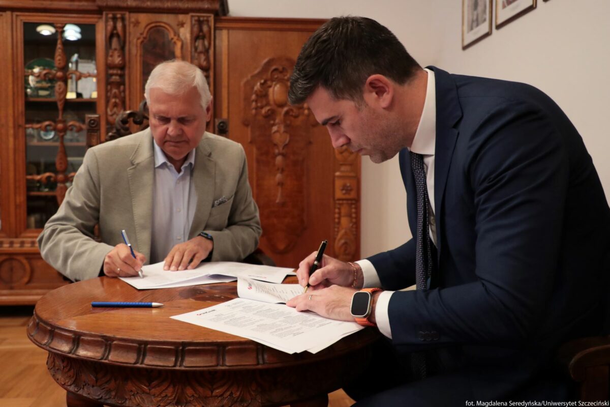Dwaj mężczyźni siedzący przy okrągłym, rzeźbionym, drewnianym stole i podpisujący dokumenty. Po lewej stronie siedzi starszy mężczyzna o siwych włosach i jasnej karnacji, ubrany w jasnoszarą marynarkę i błękitną koszulę. Skupia się na podpisywaniu kartek leżących przed nim. Po prawej stronie siedzi młodszy mężczyzna o ciemnych włosach i jasnej karnacji, ubrany w granatowy garnitur, białą koszulę i krawat. Na lewym nadgarstku ma pomarańczowy zegarek z prostokątną tarczą. On również podpisuje dokument. Na stole leżą rozłożone wydrukowane strony z tekstem oraz niebieski długopis. W tle stoi duża, ozdobna, drewniana szafa z rzeźbieniami i przeszklonymi półkami, na których znajdują się różne przedmioty. Na ścianie z prawej strony wiszą małe, oprawione w ramki obrazki lub zdjęcia.