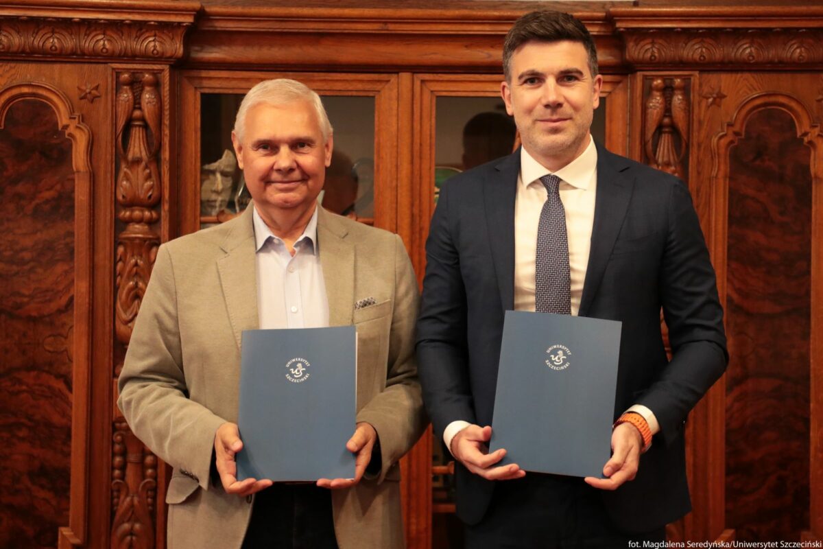Dwóch mężczyzn stojących obok siebie przed ozdobnym, drewnianym regałem. Po lewej stronie starszy mężczyzna o siwych włosach i jasnej karnacji ma na sobie jasnoszarą marynarkę, błękitną koszulę i trzyma przed sobą niebieską teczkę. Po prawej stronie młodszy mężczyzna o ciemnych włosach i jasnej karnacji jest ubrany w ciemnogranatowy garnitur, białą koszulę i krawat w drobny wzór; również trzyma przed sobą niebieską teczkę. Na teczkach widnieje białe okrągłe logo z napisem „Uniwersytet Szczeciński” i symbolem mewy pośrodku. Tło stanowi masywne, ciemnobrązowe, rzeźbione drewno z przeszklonym środkowym segmentem mebla.