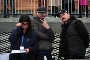 Od lewej: Beata Mikołajewska-Wieczorek – dyrektorka Centrum Edukacji Medialnej i Interaktywności z członkami jej zespołu – dwojgiem mężczyzn. Mężczyzna w środku pokazuje znak Victorii skierowany zamkniętą dłonią w swoją stronę.