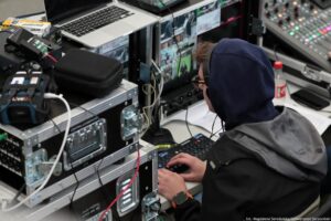 Ujęcie od góry. Mężczyzna w słuchawkach i kapturze siedzący tyłem do kadru realizuje transmisję wydarzenia. Przed nim ekrany, klawiatura, obok konsola transmisyjne z przyciskami i skrzynie na sprzęt.