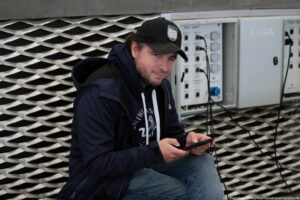 Członek ekipy Uniwizji – Telewizji Uniwersytetu Szczecińskiego w granatowej bluzie z białym logiem uczelni siedzi i trzyma telefon.