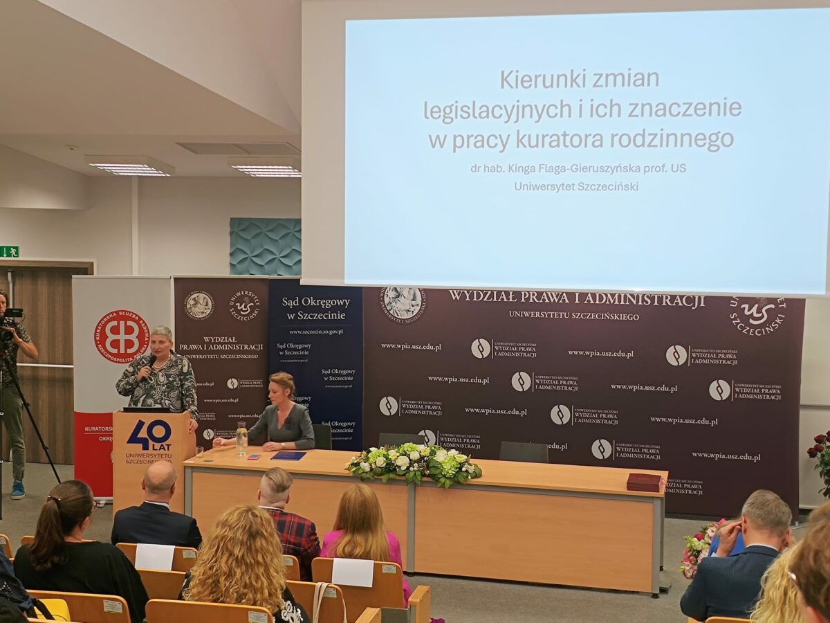 Sala wykładowa ze slajdem wyświetlonym na dużym ekranie. Na slajdzie jest tekst: “Kierunki zmian legislacyjnych i ich znaczenie w pracy kuratora rodzinnego dr hab. Kinga Flaga-Gieruszyńska prof. US Uniwersytet Szczeciński” Po lewej stoi kobieta z mikrofonem przy mównicy, a obok przy stole siedzi druga kobieta. Z przodu jest długi stół z bukietem kwiatów na środku. W tle znajdują się banery z logotypami i napisami, m.in. “WYDZIAŁ PRAWA I ADMINISTRACJI UNIWERSYTETU SZCZECIŃSKIEGO”, “Sąd Okręgowy w Szczecinie” oraz adresy “www.wpia.usz.edu.pl” i “www.szczecin.so.gov.pl”. Na mównicy widać duży napis “40 LAT” oraz “Uniwersytetu Szczecińskiego”. Na dole zdjęcia siedzi publiczność na krzesłach, widoczna od tyłu.