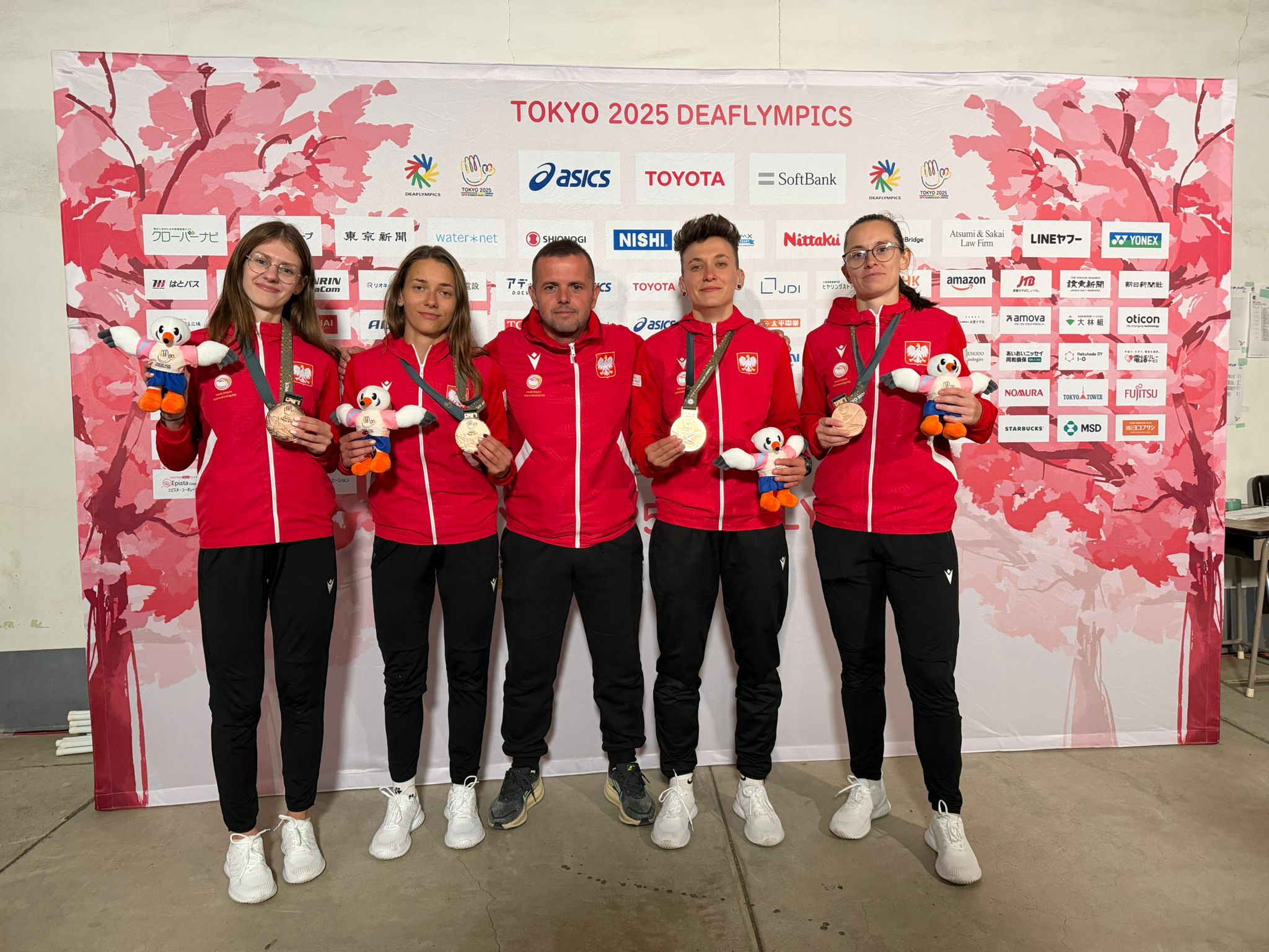 Grupa pięciu osób w czerwonych dresach z godłem Polski, stojąca na tle ścianki sponsorskiej z napisem „TOKYO 2025 DEAFLYMPICS”. Cztery kobiety, trzymając medale na wstążkach oraz pluszowe maskotki w biało-niebiesko-pomarańczowych kolorach i mężczyzna pozują do zdjęcia. Medale wyglądają na jasne, metaliczne krążki. Wszyscy mają czarne spodnie dresowe i białe buty sportowe; jedna osoba pośrodku ma ciemniejsze buty. Dwie osoby (pierwsza z lewej i pierwsza z prawej) mają okulary. Tło jest różowo-białe, z motywem kwiatów wiśni i wieloma logotypami sponsorów.
