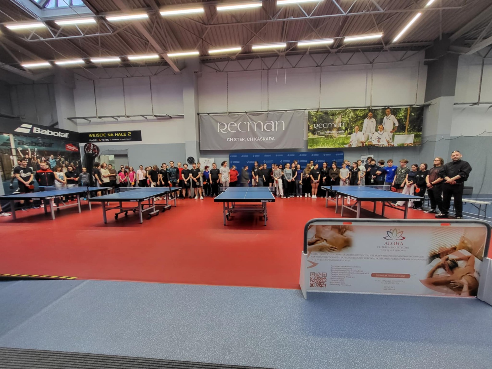 Duża hala sportowa z czerwonym podłożem i kilkoma stołami do tenisa stołowego ustawionymi na środku. W tle, wzdłuż ściany, stoi duża grupa ludzi (młodzież i dorośli) ustawiona w szeregu, jak do wspólnego zdjęcia.