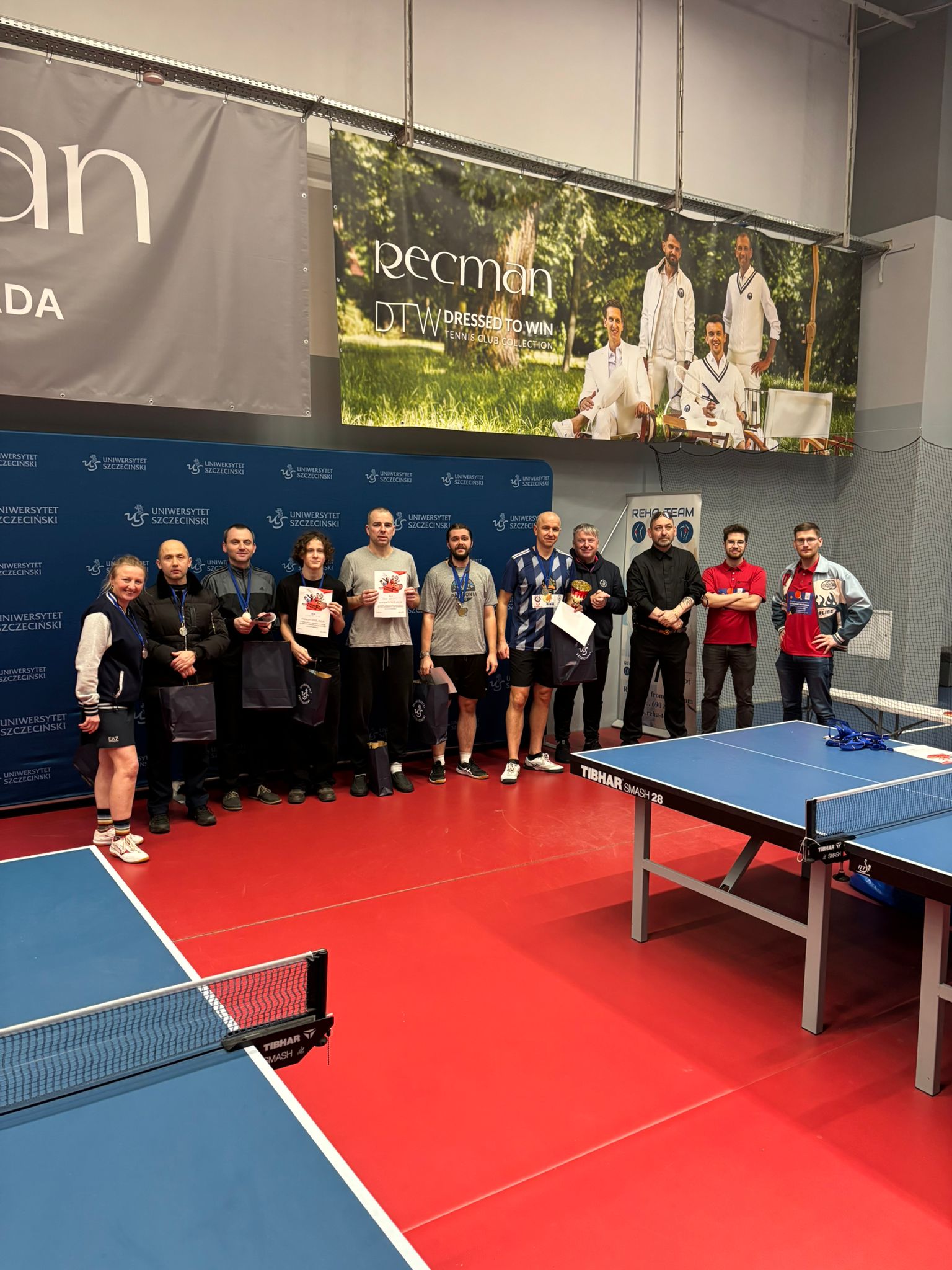 Grupa kilkunastu osób pozujących do wspólnego zdjęcia w hali do tenisa stołowego. Osoby stoją w jednym rzędzie na czerwonej podłodze, część trzyma dyplomy, medale i torby; większość to mężczyźni, po lewej stronie stoi jedna kobieta w sportowym stroju. W tle jest granatowa ścianka z powtarzającym się napisem „UNIWERSYTET SZCZECIŃSKI” i logo uczelni. Nad grupą wisi duży baner reklamowy z napisem „Peecman”, „DTW DRESSED TO WIN” oraz mniejszym tekstem „TENNIS CLUB COLLECTION”; na banerze są cztery osoby w białych strojach na tle zielonego parku.