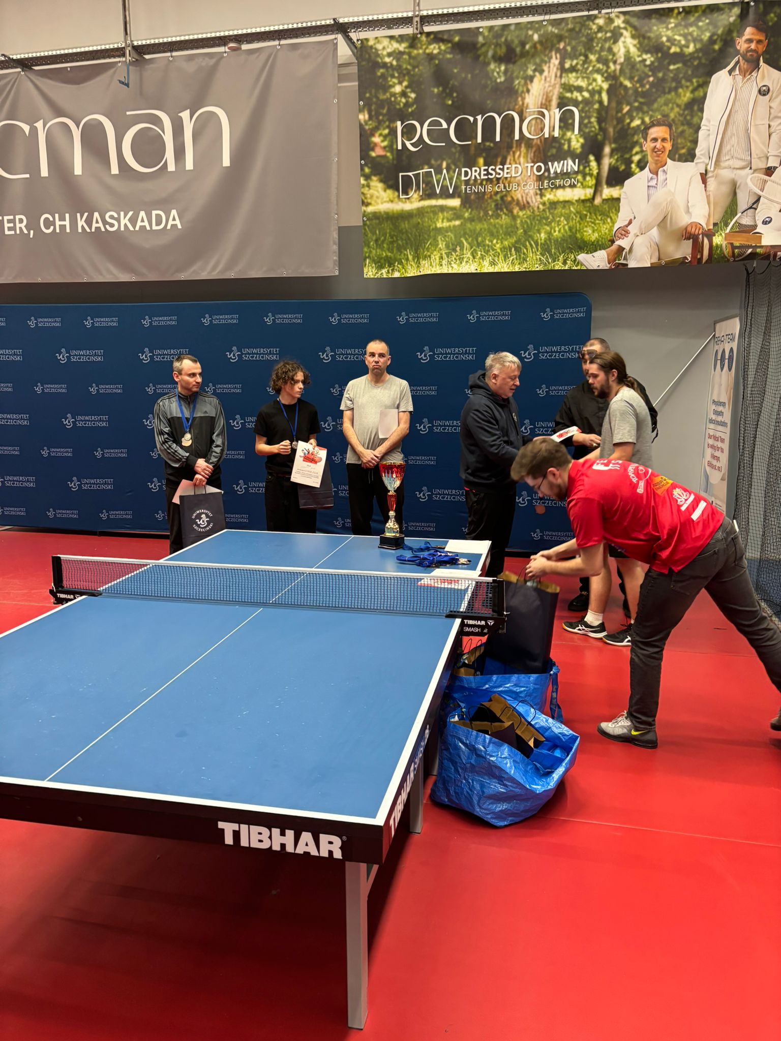 Stół do tenisa stołowego w kolorze niebieskim z siatką pośrodku, stojący na czerwonej podłodze sportowej. Na krawędzi stołu widoczny jest napis „TIBHAR”. W tle stoi grupa osób podczas ceremonii wręczania nagród. Po lewej stronie stoi mężczyzna z medalem na szyi, trzymający torbę i dokument. Obok jest młoda osoba z kręconymi włosami, trzymająca dyplom i torbę. Dalej stoi mężczyzna w jasnej koszulce z dokumentem w ręku. Na stole, bliżej środka, stoi duży puchar, a obok leżą medale na niebieskich wstążkach. Po prawej stronie dwie osoby rozmawiają przy stole; jedna z nich trzyma coś w rękach (prawdopodobnie dokument lub nagrodę). Z przodu po prawej widać osobę w czerwonej koszulce pochyloną nad torbami; na podłodze stoją duże torby (jedna niebieska, jedna ciemna).