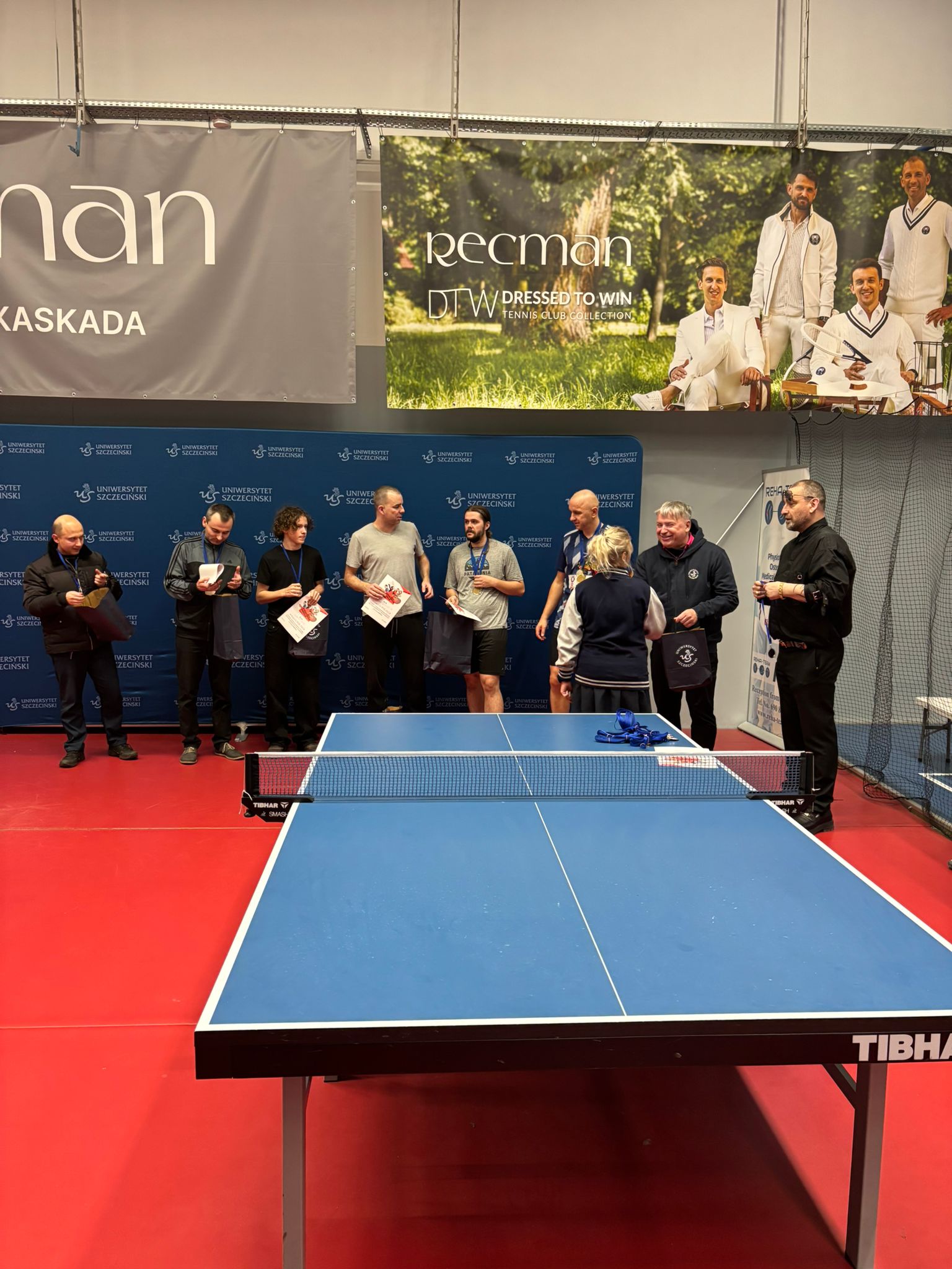 Niebieski stół do tenisa stołowego z białymi liniami i siatką, ustawiony na czerwonej podłodze w hali. Na blacie przy siatce leży kilka niebieskich smyczy lub tasiemek oraz biało-czerwony przedmiot przypominający kartkę albo małą tabliczkę.