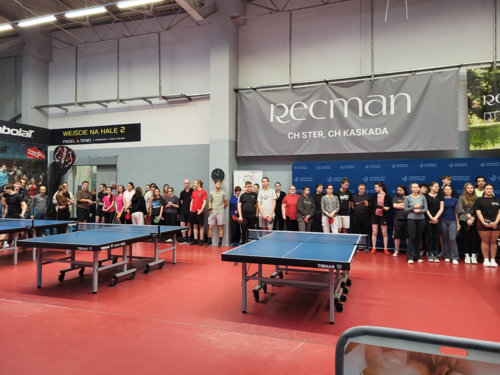 Hala sportowa z czerwonym podłożem i dwoma stołami do tenisa stołowego na pierwszym planie. Stoły są niebieskie, z siatkami, ustawione równolegle, z widocznymi kółkami w podstawach. Wzdłuż tylnej ściany stoi duża grupa ludzi ustawionych w szeregu, jak do zdjęcia grupowego lub przed rozpoczęciem wydarzenia. Osoby są w strojach sportowych i codziennych; część trzyma w dłoniach rakietki do ping-ponga.