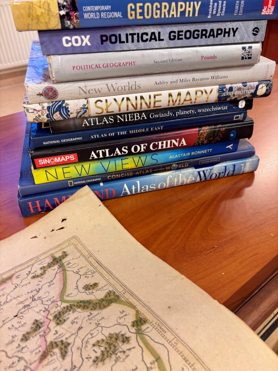 W lewym dolnym rogu archiwalna mapa. W tle stos książek dotyczących geografii i atlasów.