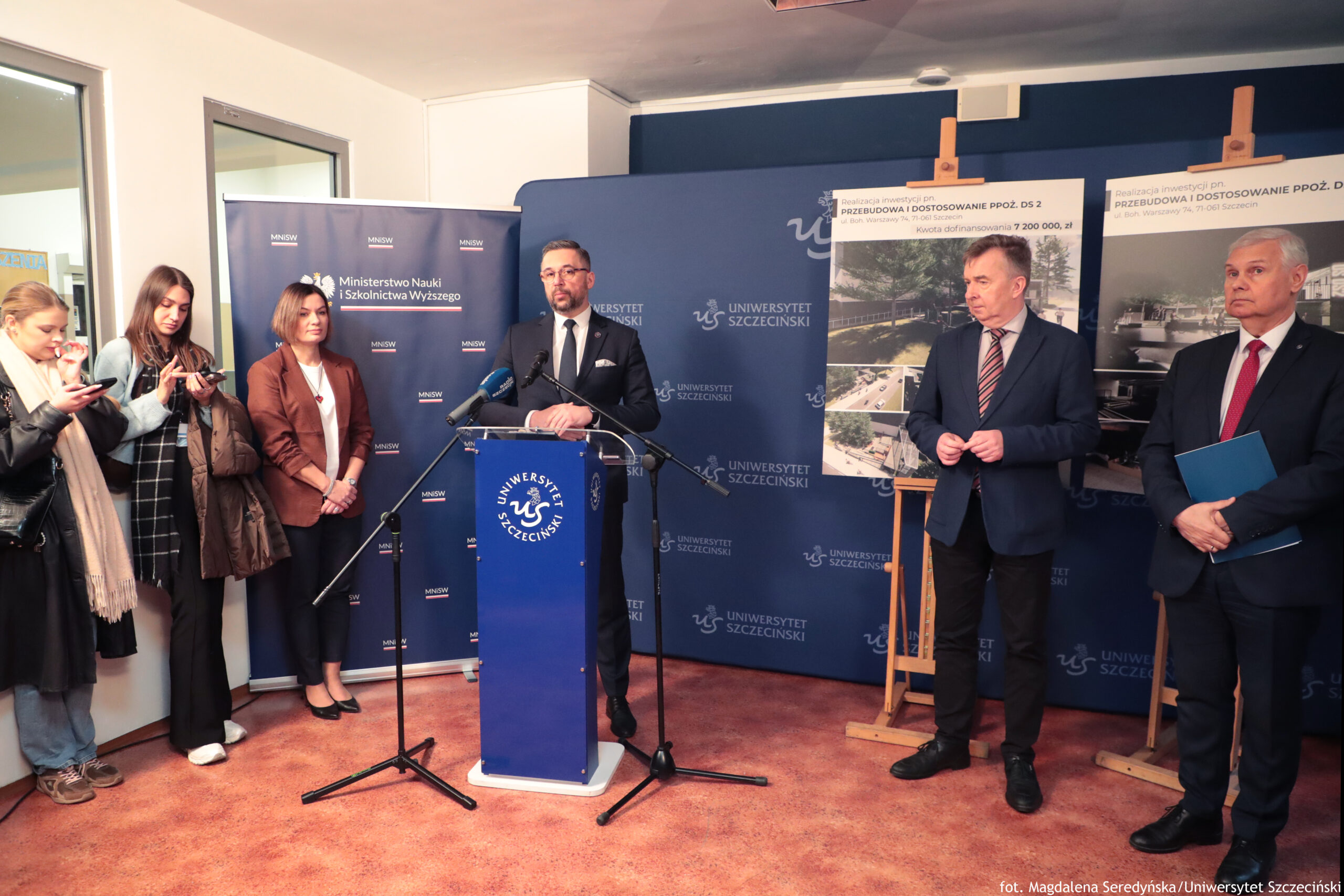 Konferencja prasowa w pomieszczeniu, z mównicą z logo „Uniwersytet Szczeciński” na pierwszym planie. Na środku stoi mężczyzna w garniturze, przy mównicy z kilkoma mikrofonami na statywach, przemawia do zebranych. Za nim i obok niego znajdują się granatowe ścianki z powtarzającymi się napisami „Uniwersytet Szczeciński” oraz baner „Ministerstwo Nauki i Szkolnictwa Wyższego” (z małymi skrótami MNiSW). Po lewej stronie stoi grupa czterech kobiet; dwie z nich trzymają telefony w dłoniach, jedna ma jasny szalik, jedna ma długie ciemne włosy. Obok nich stoi kobieta w brązowej marynarce i białej bluzce, z rękami złożonymi z przodu, zwrócona w stronę mówiącego. Po prawej stronie stoją dwaj mężczyźni w garniturach. Jeden, bliżej środka, ma ciemny garnitur i krawat w pasy, trzyma dłonie razem na wysokości brzucha i jest zwrócony w stronę mównicy. Drugi, skrajnie po prawej, ma siwe włosy, ciemny garnitur i czerwony krawat; trzyma niebieską teczkę. Za nimi stoją dwie plansze na drewnianych sztalugach z nagłówkami w języku polskim, zaczynającymi się od „PRZEBUDOWA I DOSTOSOWANIE…”, oraz zdjęciami (m.in. ujęcia budynków i infrastruktury). Na dole zdjęcia widoczny podpis: „fot. Magdalena Seredyńska/Uniwersytet Szczeciński”.