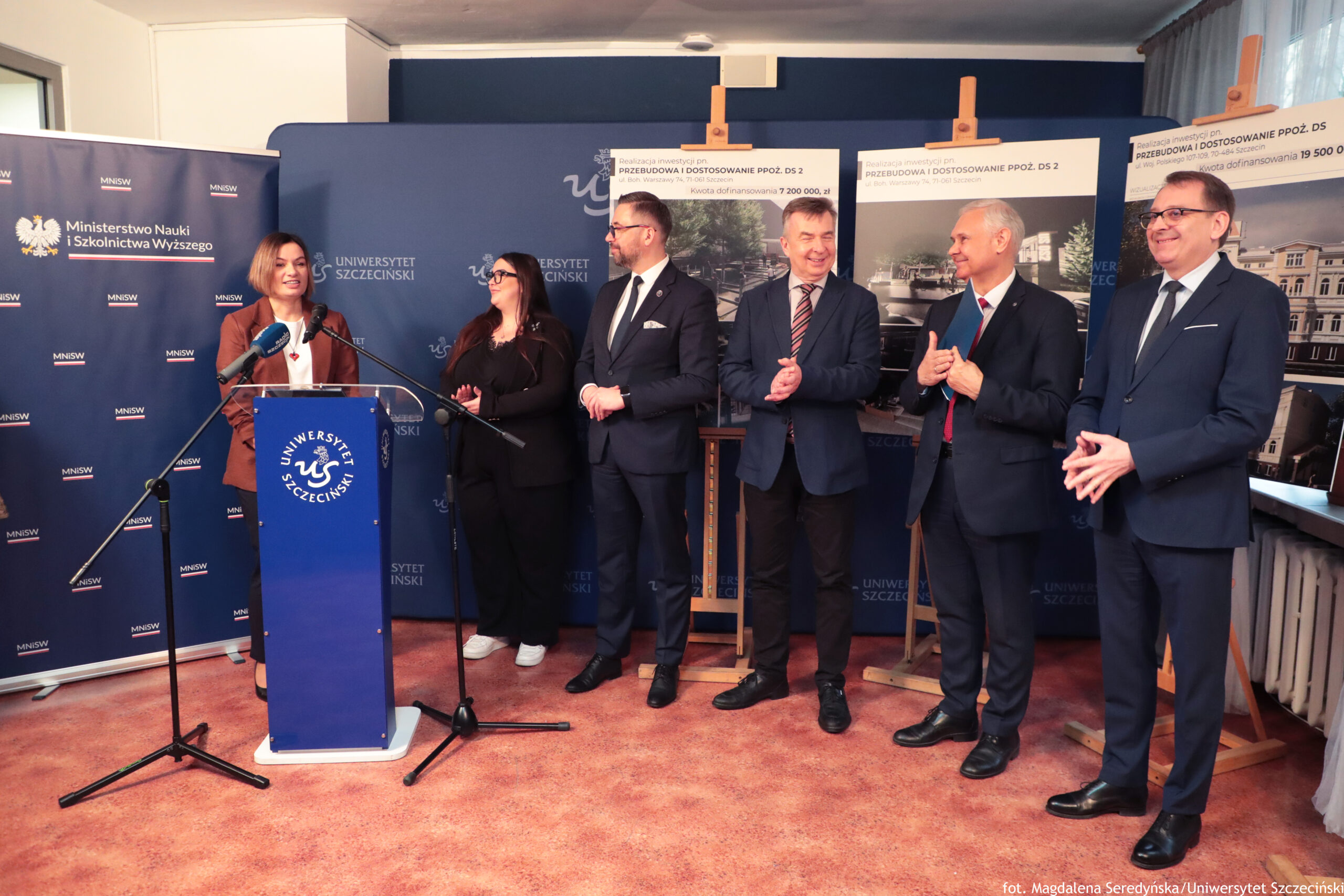 Grupa sześciu osób stojących na sali konferencyjnej przy mównicy z logo Uniwersytetu Szczecińskiego. Po lewej stronie kobieta w brązowej marynarce stoi przy niebieskiej mównicy i mówi do mikrofonów na statywach. Obok niej stoi kobieta w czarnym stroju, a dalej czterech mężczyzn w garniturach; kilku z nich ma dłonie złożone jak do oklasków i uśmiecha się. W tle są granatowe ścianki i roll-upy z napisami „Ministerstwo Nauki i Szkolnictwa Wyższego” oraz „Uniwersytet Szczeciński”. Po prawej stronie stoją duże plansze na sztalugach z tytułem „PRZEBUDOWA I DOSTOSOWANIE PPOŻ. DS 2” oraz widocznymi kwotami dofinansowania. W prawym dolnym rogu jest podpis: „fot. Magdalena Seredyńska / Uniwersytet Szczeciński”.