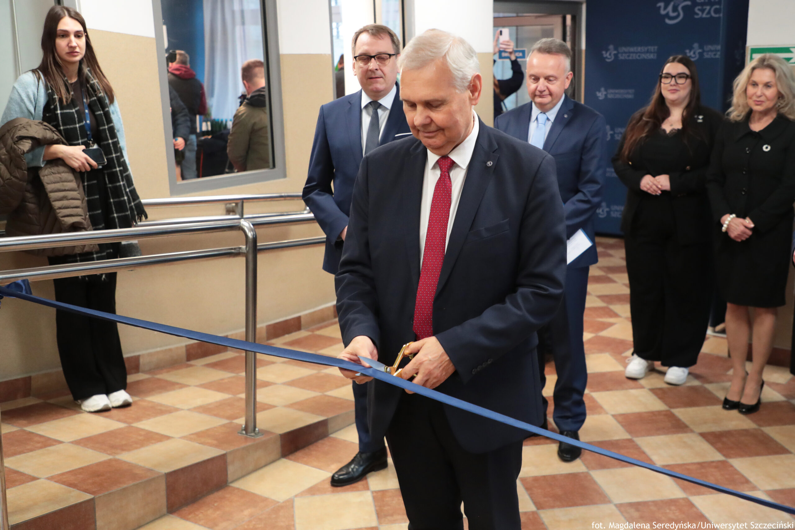 Ceremonia przecięcia wstęgi w budynku, z mężczyzną w garniturze na pierwszym planie trzymającym nożyczki przy niebieskiej wstędze. Mężczyzna na pierwszym planie ma siwe włosy, ciemny granatowy garnitur, białą koszulę i czerwoną krawat. Wstęga biegnie ukośnie przez kadr, mniej więcej na wysokości jego rąk. Za nim stoją dwaj mężczyźni w garniturach; jeden w okularach, drugi z krótkimi włosami. Po prawej stronie kadru stoją dwie kobiety w czarnych ubraniach; jedna ma długie ciemne włosy i okulary, druga jasne blond włosy do ramion. Po lewej stronie stoi młoda kobieta z długimi brązowymi włosami, w jasnym płaszczu i czarno-białym szaliku, trzyma w rękach kurtkę. W tle widać granatową ściankę z powtarzającym się napisem „UNIWERSYTET SZCZECIŃSKI” i skrótem „US”. Podłoga jest w brązowo-beżową kratę z płytek, a obok znajdują się metalowe poręcze i stopnie. W prawym dolnym rogu jest podpis: „fot. Magdalena Seredyńska/Uniwersytet Szczeciński”.