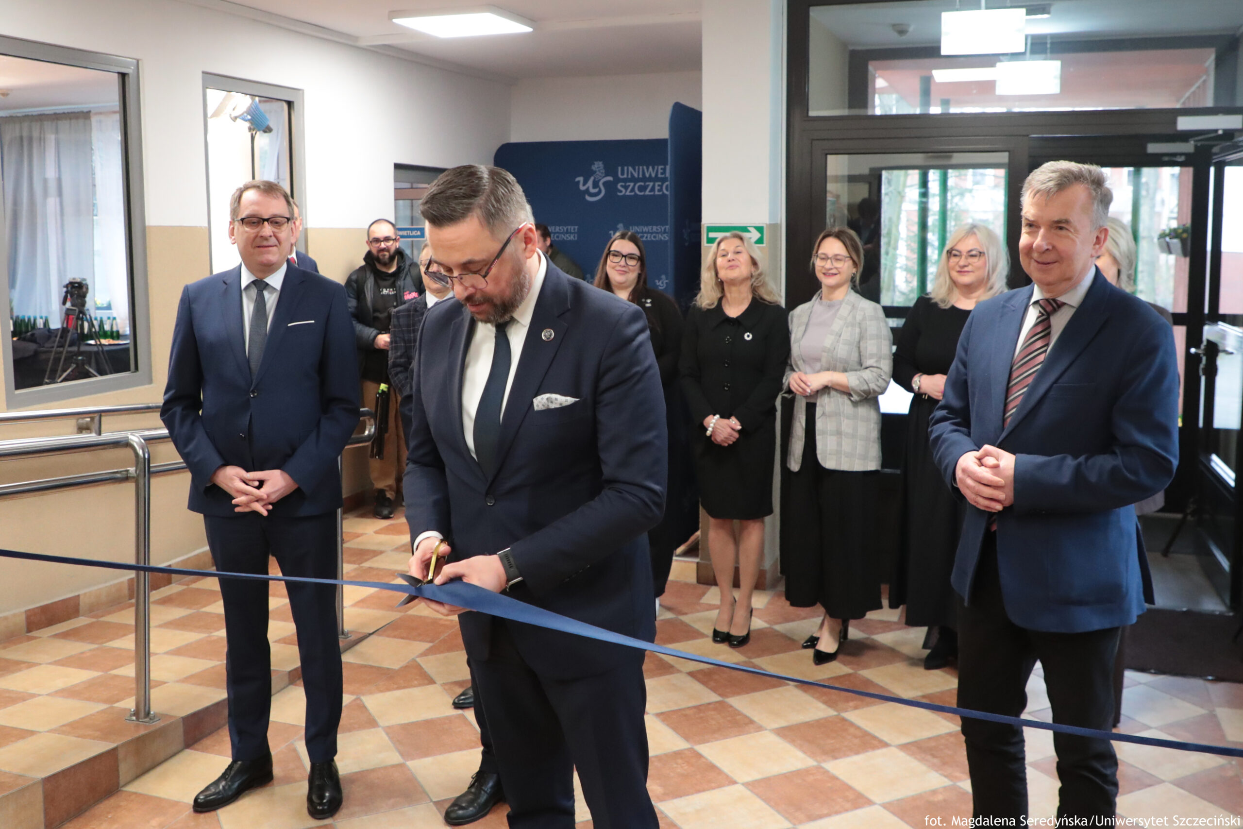 Grupa osób w holu budynku podczas uroczystego przecięcia wstęgi. Na pierwszym planie stoi mężczyzna w granatowym garniturze, białej koszuli i ciemnym krawacie, z okularami i przystrzyżoną brodą; trzyma nożyczki przy napiętej, ciemnoniebieskiej wstędze rozciągniętej na wysokości pasa. Po lewej stronie stoi inny mężczyzna w granatowym garniturze, białej koszuli i jasnym krawacie, w okularach, z dłońmi złożonymi z przodu. Po prawej stronie stoi mężczyzna w niebieskiej marynarce, jasnej koszuli i paskowanym krawacie, z dłońmi złożonymi, lekko uśmiechnięty. W tle stoi kilka osób, głównie kobiety w formalnych strojach (czarne sukienki, jasna marynarka w kratę), część w okularach. Widać wejście z przeszklonymi drzwiami oraz elementy wyposażenia holu: poręcz, podest i posadzkę w beżowo-brązową kratę. Na granatowym tle pośrodku jest widoczny napis „UNIWERSYTET SZCZECIŃSKI” oraz logo „US”. W prawym dolnym rogu znajduje się podpis: „fot. Magdalena Seredyńska/Uniwersytet Szczeciński”.