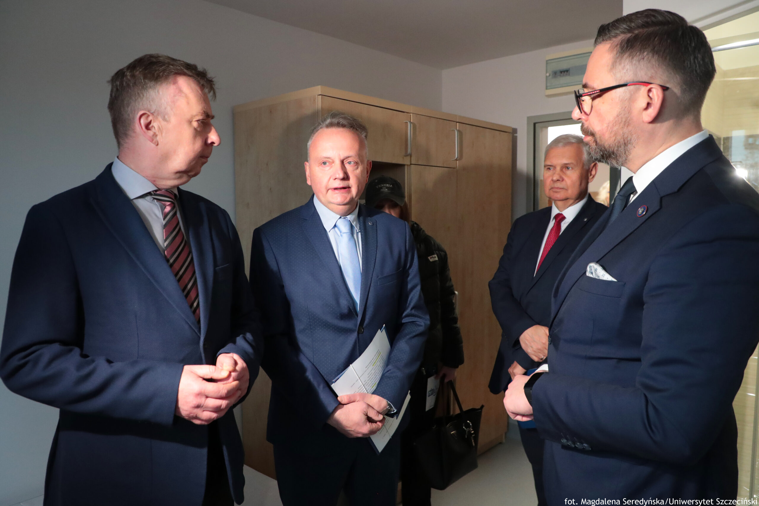 Grupa czterech mężczyzn w garniturach stojących w pomieszczeniu biurowym i rozmawiających. Kadr pokazuje ich od pasa w górę, w półprofilach, w dwóch małych grupach zwróconych do siebie. Po lewej stoi mężczyzna w ciemnogranatowym garniturze, białej koszuli i krawacie w skośne pasy (bordowo-szare). Ma krótkie, jasnobrązowe włosy i jest zwrócony twarzą w stronę środka kadru. W centrum stoi mężczyzna w granatowym garniturze i jasnoniebieskim krawacie, z krótkimi siwiejącymi włosami. Trzyma w rękach kilka kartek dokumentów. Po prawej, najbliżej aparatu, stoi mężczyzna w ciemnym garniturze, białej koszuli i ciemnym krawacie, w okularach, z krótkimi ciemnymi włosami i przystrzyżoną brodą. W brustaszy ma jasną poszetkę; na klapie marynarki widać okrągłą przypinkę. W tle, między mężczyznami po prawej, stoi starszy mężczyzna w granatowym garniturze i czerwonym krawacie, z krótkimi siwymi włosami. Za centralną postacią widać jeszcze częściowo zasłoniętą osobę w ciemnej kurtce i czapce. W tle znajduje się jasna, drewniana szafa z kilkoma drzwiczkami i metalowymi uchwytami oraz przeszklone drzwi/okno po prawej. Na dole po prawej jest podpis: „fot. Magdalena Seredyńska / Uniwersytet Szczeciński”.