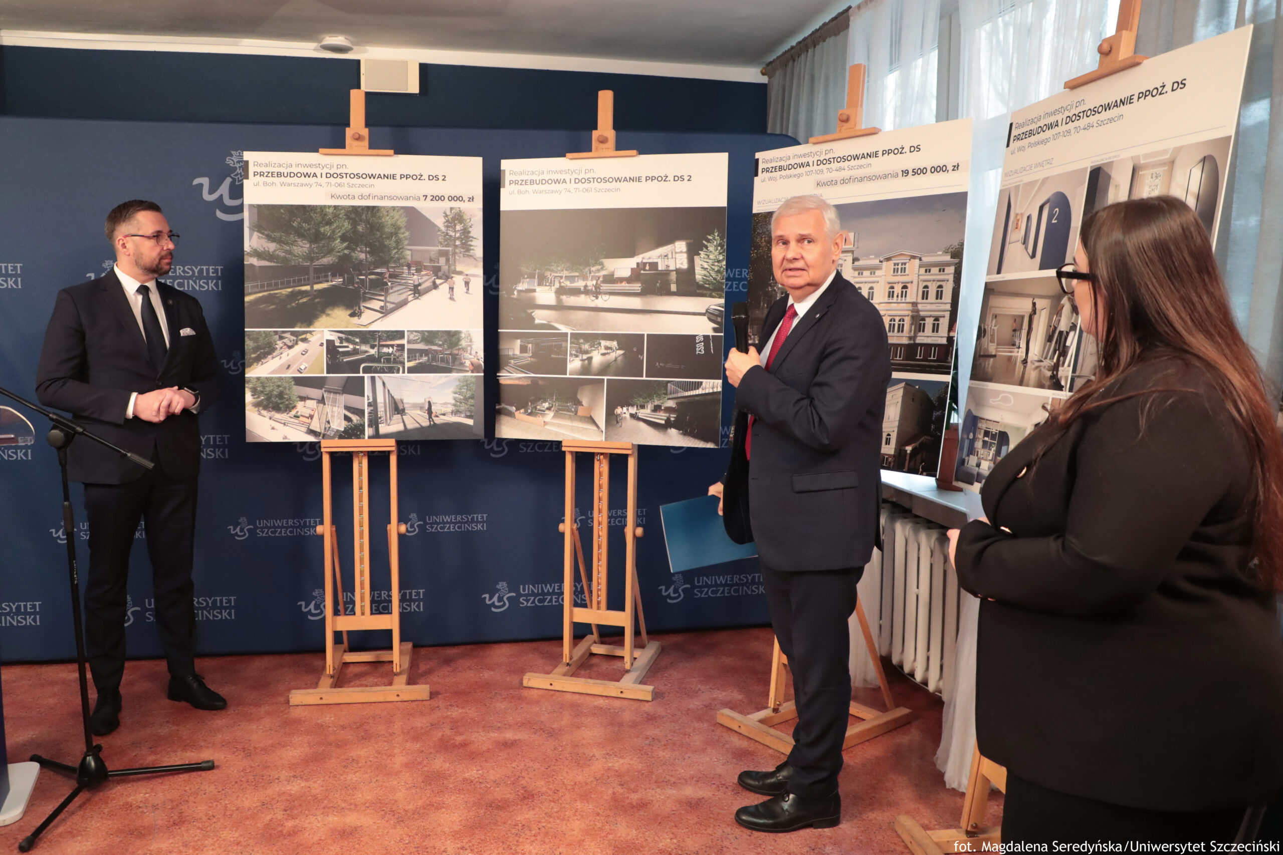 Hol z trzema osobami stojącymi przed planszami na sztalugach i granatową ścianką z powtarzającym się napisem „UNIWERSYTET SZCZECIŃSKI”. W tle po prawej stronie jest okno z jasnymi zasłonami i grzejnik pod oknem, a na podłodze czerwony dywan. Po lewej stoi mężczyzna w czarnym garniturze, białej koszuli i czarnym krawacie, w okularach. Obok niego znajduje się statyw z mikrofonem. Na środku stoi starszy mężczyzna w ciemnym garniturze z czerwonym krawatem, zwrócony przodem. W pobliżu, po prawej, stoi kobieta w czarnym stroju, z długimi włosami i okularami, ustawiona bokiem do obiektywu. Za osobami stoją trzy duże plansze na drewnianych sztalugach z wizualizacjami i zdjęciami. Na górze dwóch środkowych plansz czytelny jest tytuł: „PRZEBUDOWA I DOSTOSOWANIE PPOŻ. DS 2”. Na prawej planszy w nagłówku widać: „PRZEBUDOWA I DOSTOSOWANIE PPOŻ. DS …” (końcówka jest poza ostrością/kadrem). Na planszach są renderowane ujęcia budynku i wnętrz, w tym korytarze i schody. W prawym dolnym rogu zdjęcia jest podpis: „fot. Magdalena Seredyńska/Uniwersytet Szczeciński”.