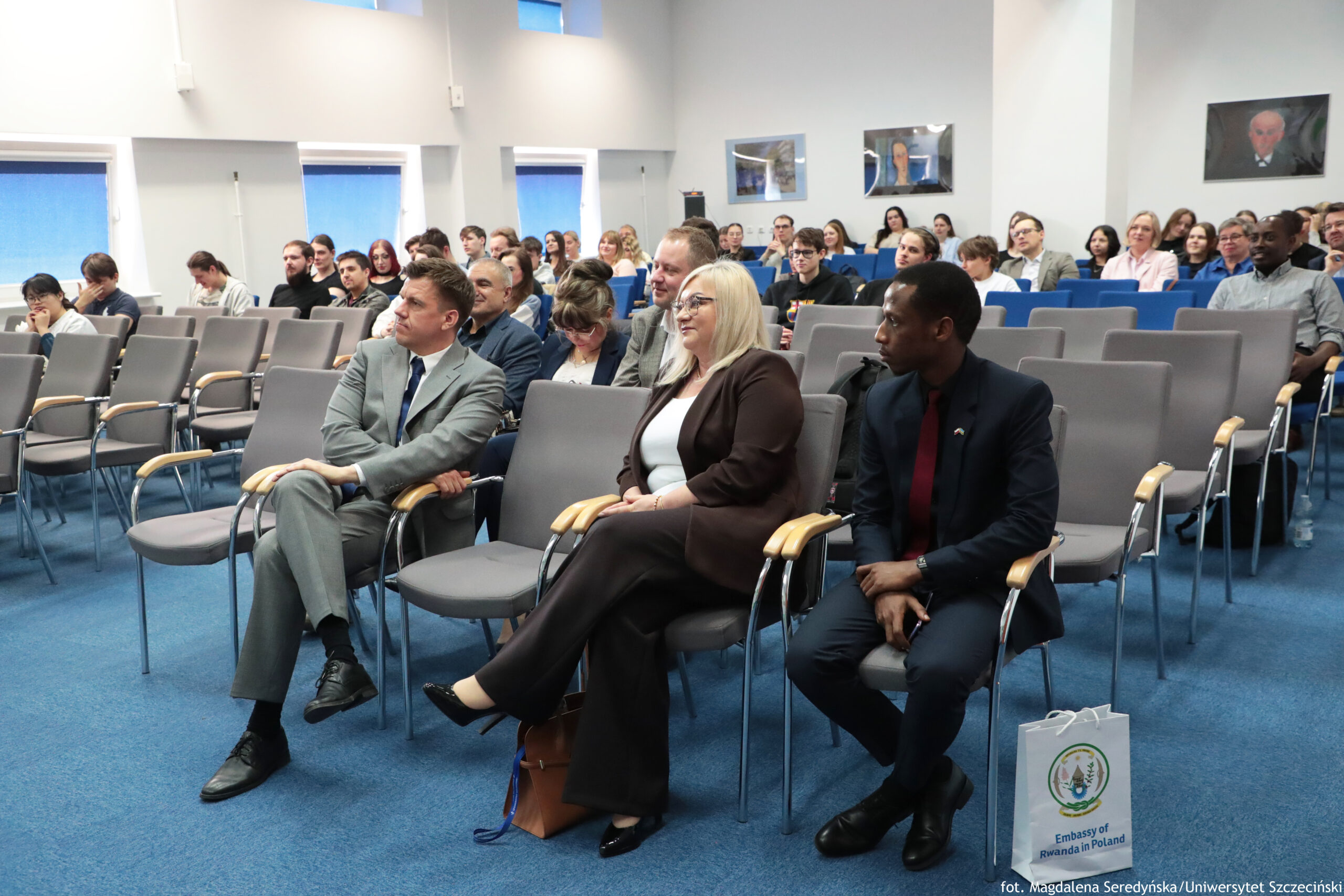 Sala konferencyjna z publicznością siedzącą na rzędach krzeseł. Na pierwszym planie, w pierwszym rzędzie, siedzą trzy osoby w formalnych strojach: mężczyzna w jasnoszarym garniturze, kobieta w ciemnym garniturze z jasnymi blond włosami i okularami oraz mężczyzna o ciemnej karnacji w granatowym garniturze z bordowym krawatem. Wokół nich widać wiele pustych krzeseł, a dalej kilkadziesiąt osób zajmujących miejsca w kolejnych rzędach. Pomieszczenie ma niebieską wykładzinę i jasne ściany. Po lewej stronie są okna z opuszczonymi roletami w niebieskim odcieniu. Na ścianie z tyłu wiszą dwa obrazy lub fotografie w ramkach oraz po prawej większy portret. Przy prawym dolnym rogu obrazu stoi biała torba z nadrukiem i czytelnym napisem „Embassy of Rwanda in Poland”. Na dole zdjęcia jest też podpis fotograficzny: „fot. Magdalena Poredyńska/Uniwersytet Szczeciński”.