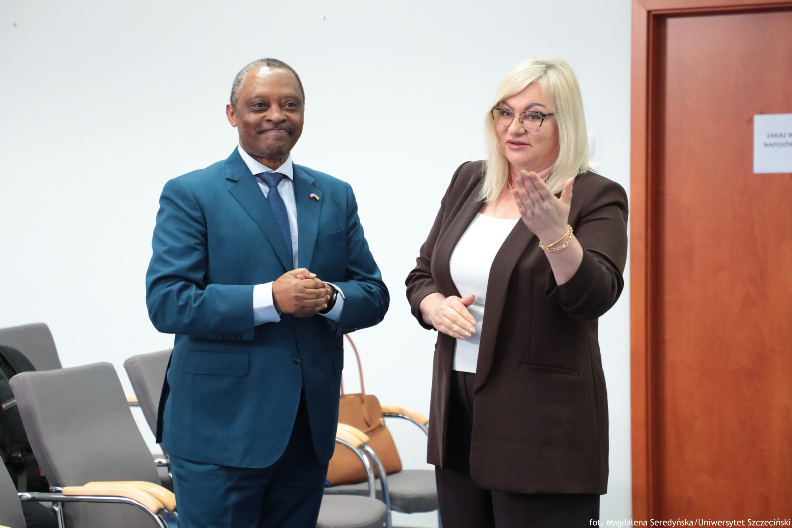 Dwie osoby stojące w pomieszczeniu biurowym na tle jasnej ściany. Po lewej stronie stoi mężczyzna o ciemnej karnacji, z bardzo krótko przyciętymi włosami, ubrany w niebieski garnitur, białą koszulę i krawat. Ma złożone dłonie na wysokości brzucha i lekki uśmiech. Na klapie marynarki widać małą przypinkę. Po prawej stronie stoi kobieta o jasnej karnacji, z prostymi, jasnymi blond włosami do ramion i w okularach. Ma na sobie ciemny (brązowy) zestaw: marynarkę i spodnie oraz białą bluzkę. Jedną dłoń trzyma uniesioną w geście, jak podczas mówienia, a drugą ma niżej, przy tułowiu. Na nadgarstku widać bransoletkę. W tle są szare krzesła z jasnymi, drewnianymi podłokietnikami oraz beżowa torebka oparta o jedno z krzeseł. Po prawej stronie jest brązowe drzwi z kartką z tekstem, którego nie da się tu jednoznacznie odczytać. W prawym dolnym rogu zdjęcia widoczny jest podpis fotograficzny: „fot. Magdalena Seredyńska/Uniwersytet Szczeciński”.
