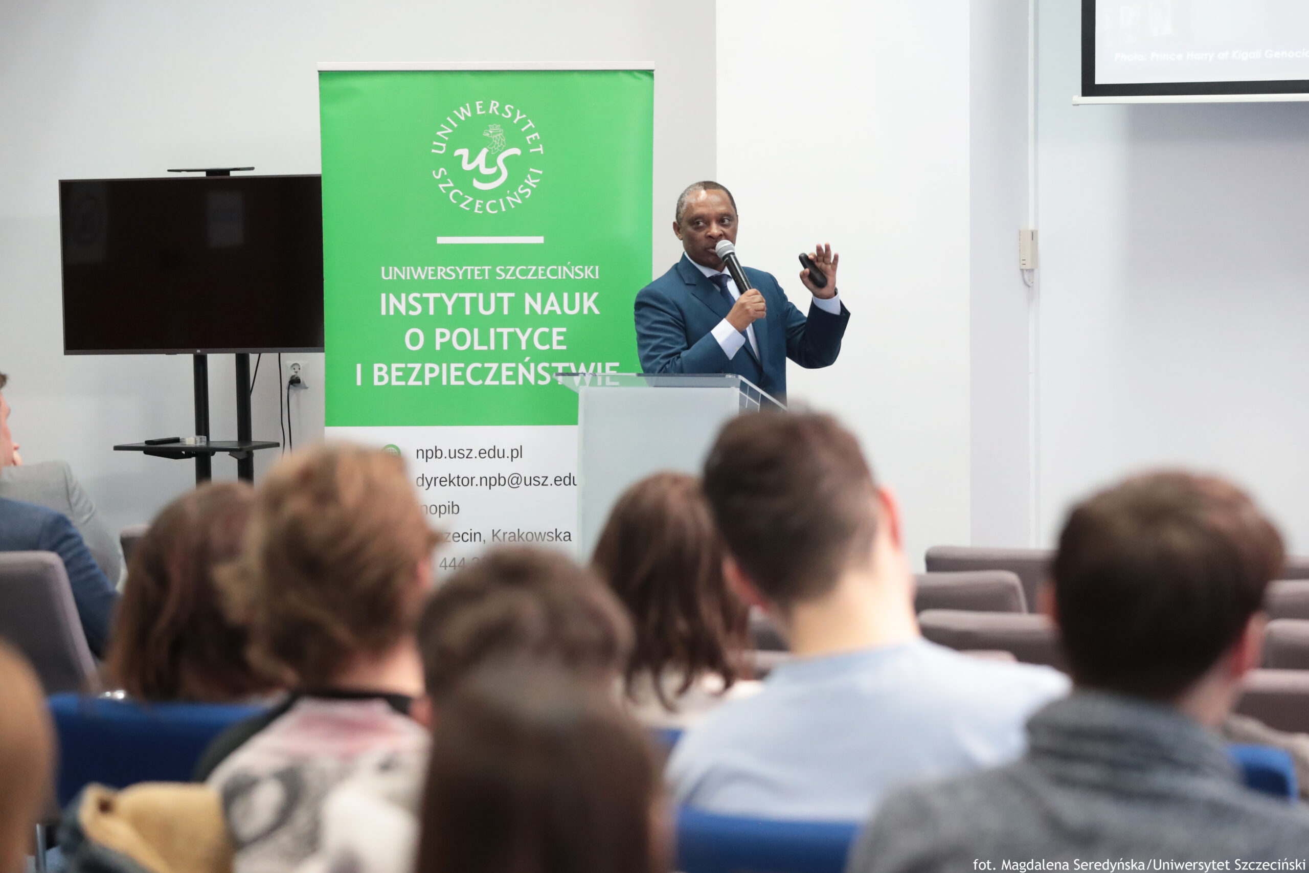 Sala konferencyjna z prelegentem stojącym przy mównicy i przemawiającym do publiczności. Na pierwszym planie widać tyły głów kilku osób siedzących na krzesłach; twarze są niewidoczne. Pośrodku, przy mównicy, stoi mężczyzna o ciemnej karnacji, w granatowym garniturze i białej koszuli. Trzyma mikrofon przy ustach, a drugą rękę ma uniesioną w geście; w dłoni jest mały, ciemny pilot lub wskaźnik. Za nim znajduje się duży zielony roll-up z logotypem „US” i napisem „UNIWERSYTET SZCZECIŃSKI” oraz „INSTYTUT NAUK O POLITYCE I BEZPIECZEŃSTWIE”. Po lewej stronie stoi duży, czarny ekran telewizora na stojaku. W prawym górnym rogu jest fragment ekranu projekcyjnego z nieczytelnym tekstem. W prawym dolnym rogu zdjęcia widnieje podpis: „fot. Magdalena Seredyńska / Uniwersytet Szczeciński”.