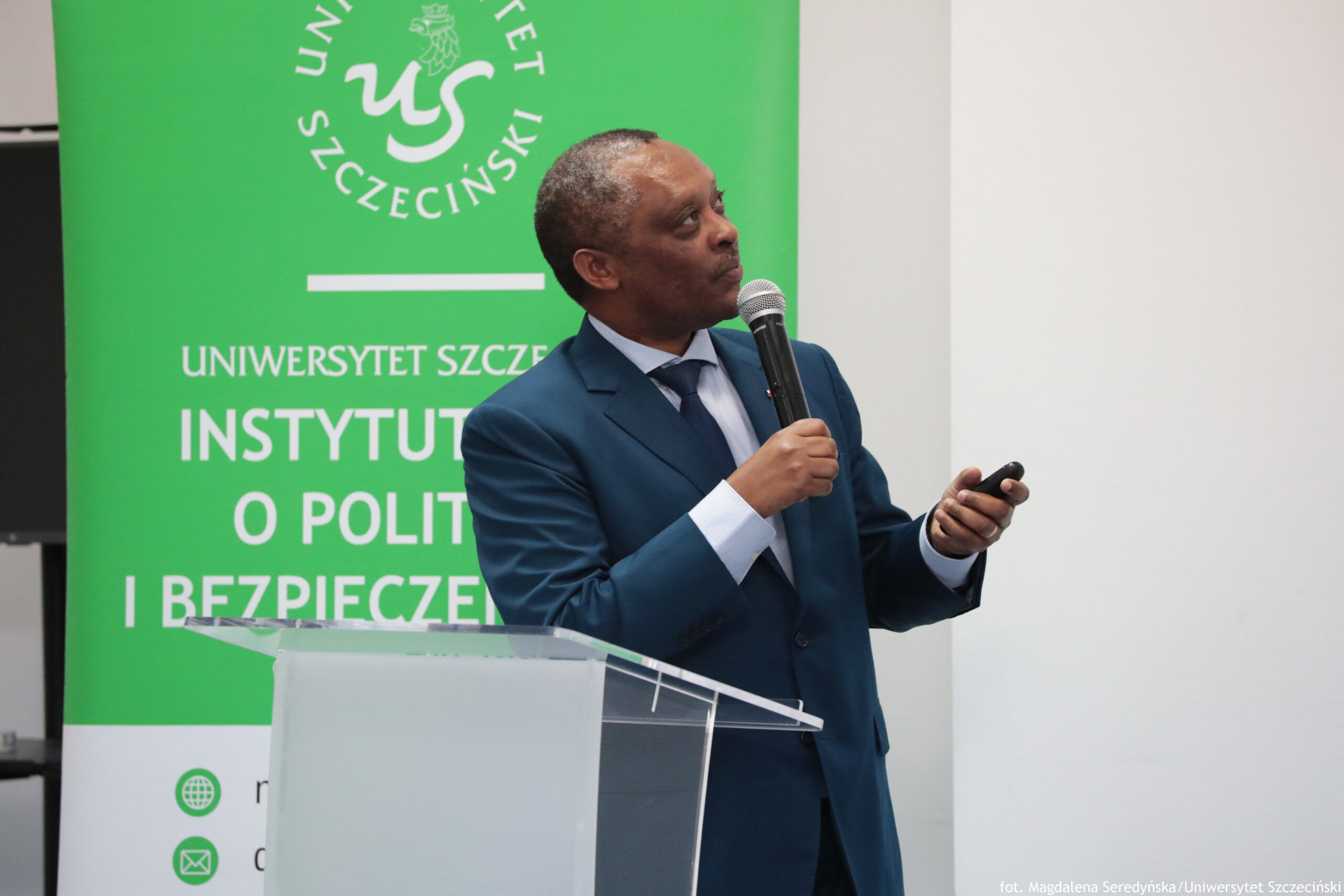 Mężczyzna w granatowym garniturze przemawia przy przezroczystej mównicy, trzymając mikrofon i pilot do prezentacji. Ma ciemną skórę, krótkie, siwiejące włosy i jest pokazany z profilu, zwrócony w prawą stronę.