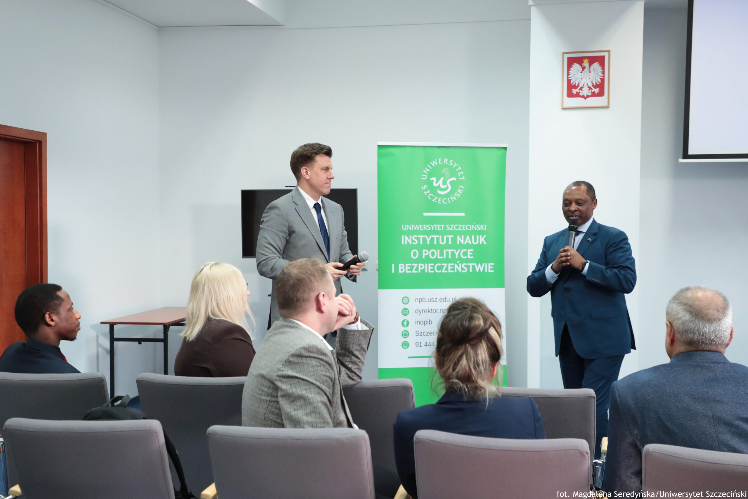 Sala konferencyjna na pierwszym planie widać rzędy szarych krzeseł i kilka osób siedzących tyłem lub bokiem; jedna osoba ma jasne, blond włosy, inna ma włosy związane w kok, widać też mężczyznę z krótkimi włosami w szarej marynarce oraz osobę o ciemnej skórze w czarnym ubraniu. Z przodu sali stoją dwaj mężczyźni w garniturach. Po lewej: mężczyzna o jasnej skórze w jasnoszarym garniturze, białej koszuli i ciemnym krawacie, trzyma w rękach mikrofon i małe urządzenie. Po prawej: mężczyzna o ciemnej skórze w granatowym garniturze, białej koszuli i ciemnym krawacie, mówi do mikrofonu, trzymając go przy ustach. Na ścianie po prawej, wysoko, wisi godło Polski (biały orzeł na czerwonym tle). W prawym górnym rogu kadru widać fragment ekranu projekcyjnego. W lewym tle jest brązowe wejście/drzwi i mały stolik przy ścianie. Na dole po prawej widnieje podpis: „fot. Magdalena Seredyńska/Uniwersytet Szczeciński”.