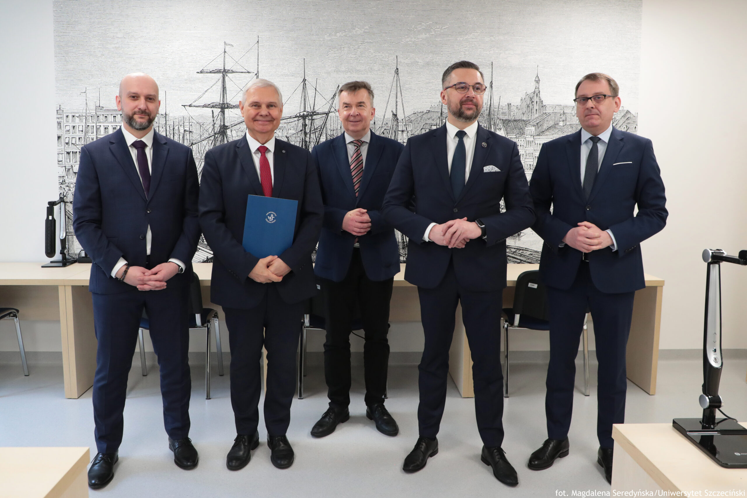 Grupa pięciu mężczyzn w garniturach stojących w pomieszczeniu biurowym na tle dużej czarno-białej grafiki przedstawiającej port z żaglowcami i zabudową miasta. Wszyscy stoją w jednym rzędzie, zwróceni przodem do aparatu, z dłońmi złożonymi z przodu; wyglądają na osoby w wieku około 40–60 lat, o jasnej karnacji. Od lewej: mężczyzna łysy z krótką brodą, w granatowym garniturze i białej koszuli; krawat w ciemnym kolorze. Drugi mężczyzna ma krótkie siwe włosy, uśmiecha się, ma czerwony krawat i trzyma niebieską teczkę z małym białym symbolem pośrodku. Trzeci mężczyzna stoi lekko z tyłu, ma krótkie brązowe włosy i krawat w ukośne pasy. Czwarty mężczyzna ma krótkie ciemne włosy, brodę i okulary; ma ciemny garnitur, białą koszulę, ciemny krawat oraz jasną poszetkę w kieszeni marynarki. Piąty mężczyzna ma krótkie jasnobrązowe włosy, okulary i granatowy garnitur z jasnym krawatem.