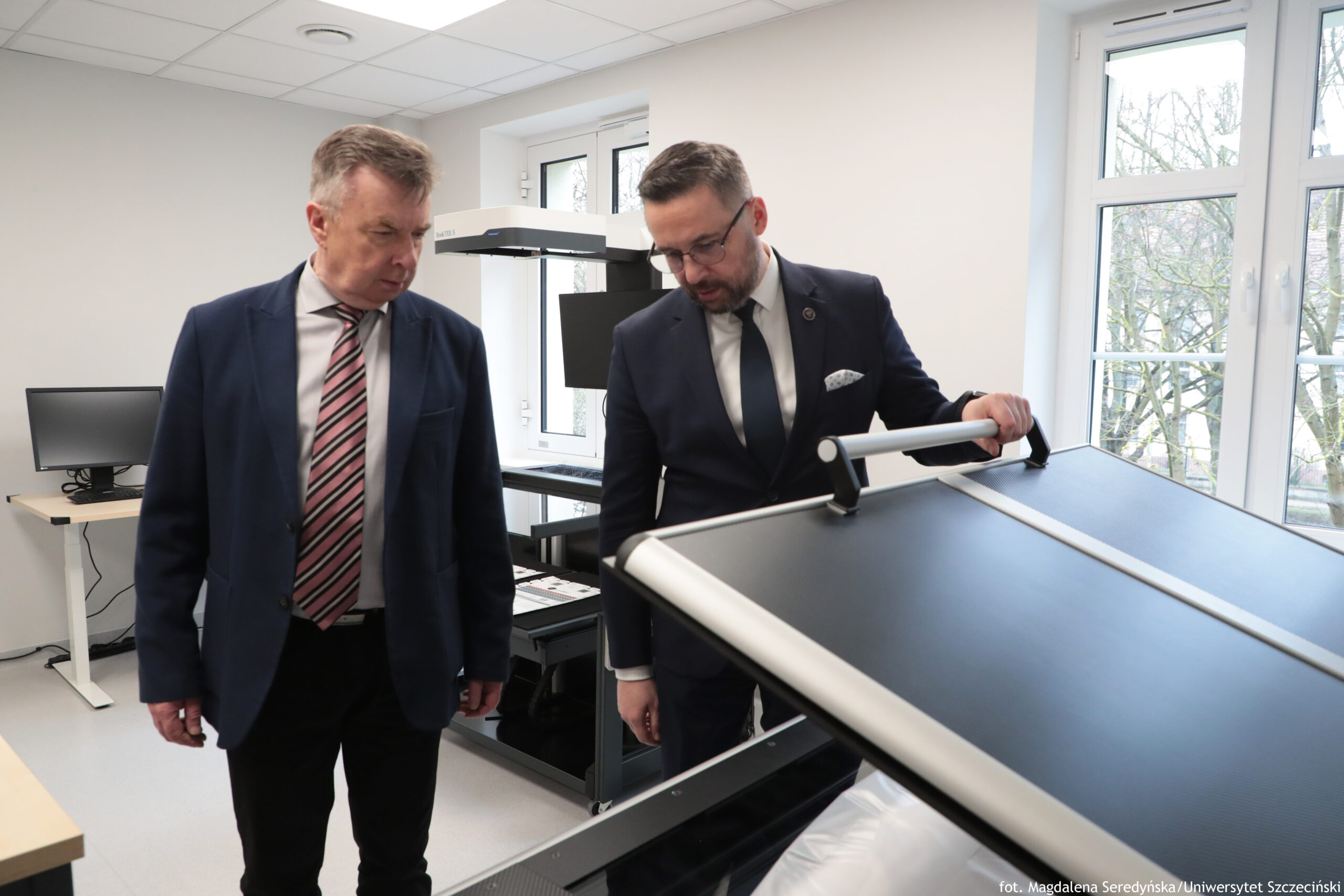 Dwóch mężczyzn w garniturach w jasnym pomieszczeniu biurowo-laboratoryjnym, stojących przy dużym urządzeniu z szerokim, czarnym pasem i metalową ramą. Mężczyzna po lewej ma siwe włosy, granatową marynarkę i krawat w różowo-szare pasy; patrzy w dół w stronę urządzenia. Mężczyzna po prawej ma krótkie ciemne włosy, brodę i okulary; ma ciemny garnitur, białą koszulę i ciemny krawat, a jedną rękę opiera na poręczy urządzenia, również kierując wzrok w dół. W tle widać biurko z monitorem, inne stanowisko z dużym ekranem oraz duże okna z widokiem na drzewa. W prawym dolnym rogu jest mały podpis: „fot. Magdalena Seredyńska/Uniwersytet Szczeciński”.